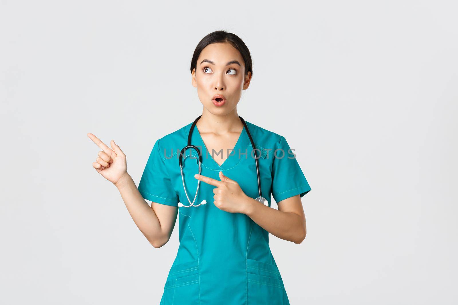 Covid-19, healthcare workers, pandemic concept. Intrigued and amazed beautiful asian female intern, nurse in scrubs or surgeon pointing and looking upper left corner, saying wow in amazement.