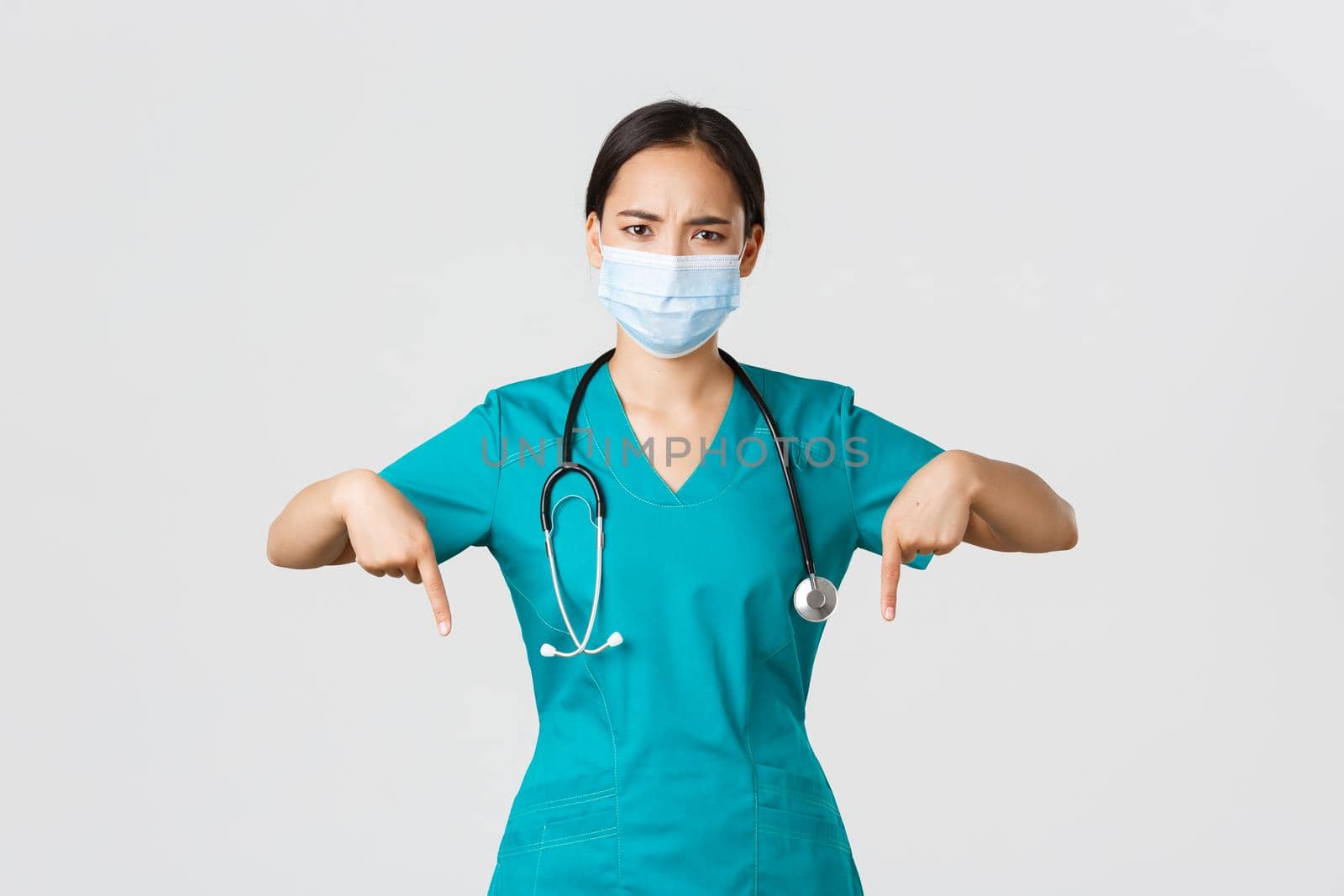 Covid-19, coronavirus disease, healthcare workers concept. Serious-looking worried asian female doctor inform patients, physician in medical mask and scrubs frowning, pointing fingers down.