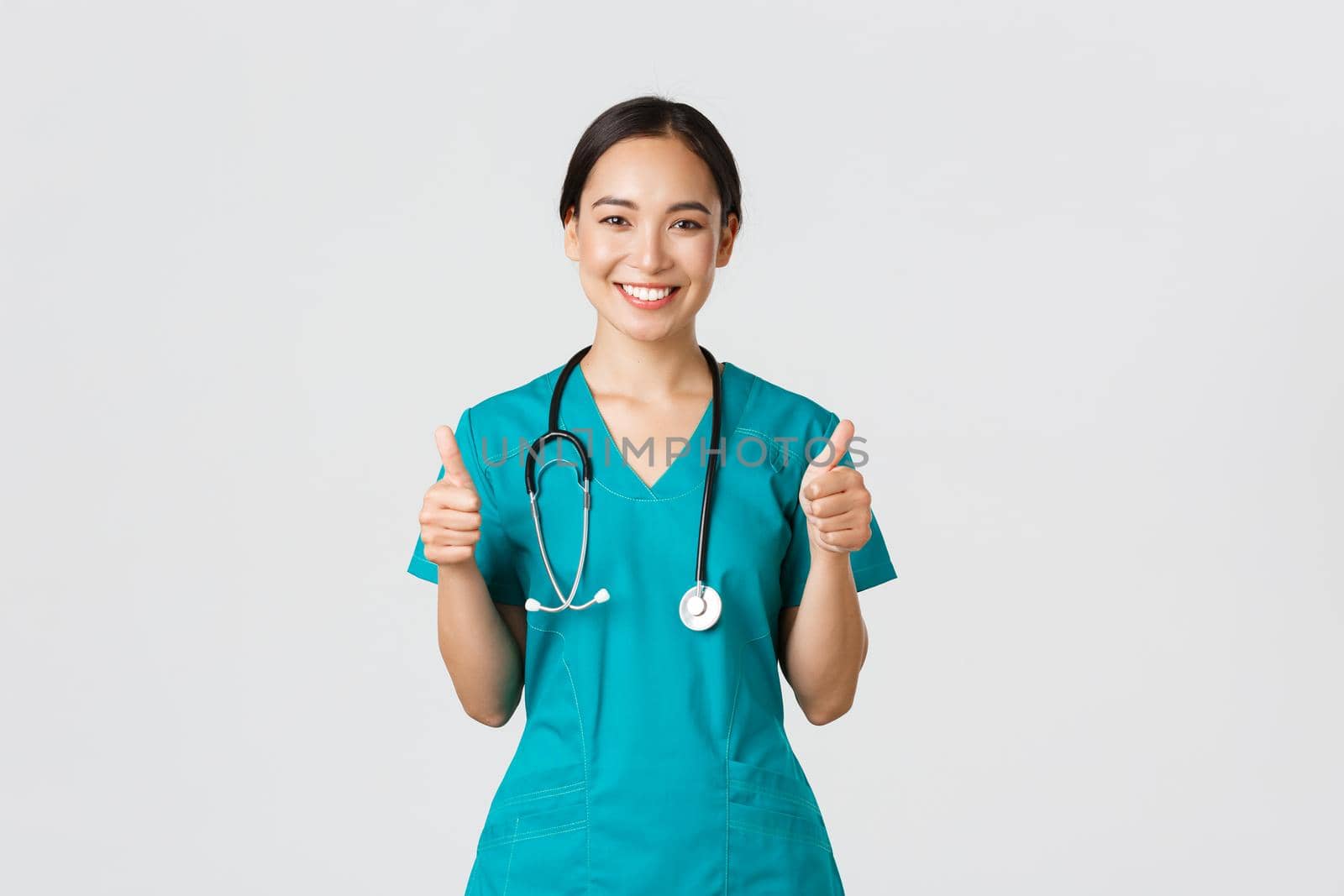 Covid-19, healthcare workers, pandemic concept. Professional confident smiling doctor, female physician in scrubs showing thumbs-up with assured expression, ensure all good, perfect or excellent.