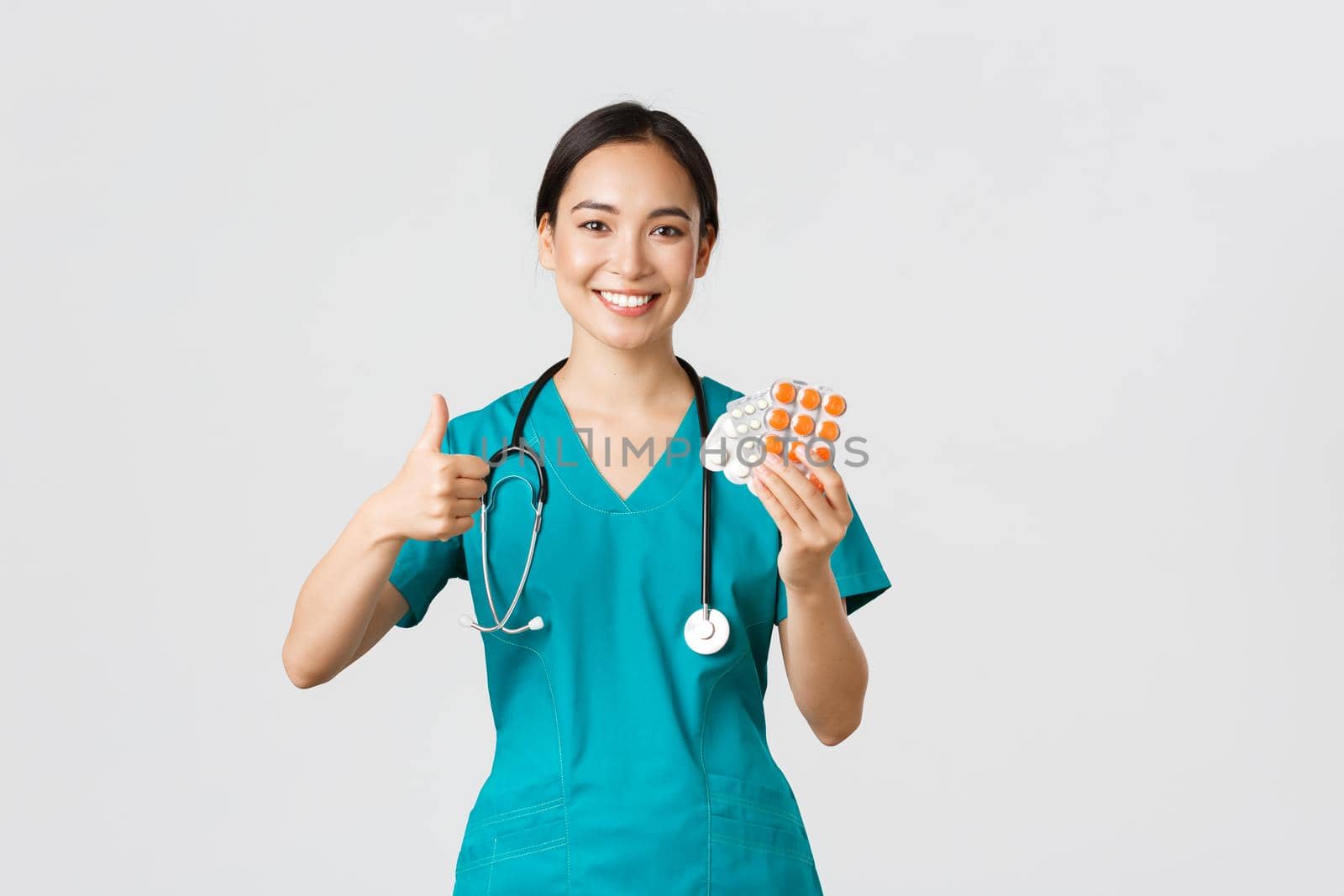 Healthcare workers, preventing virus, quarantine campaign concept. Confident smiling asian female nurse, doctor in scrubs recommend medication, show thumbs-up and pills, white background.