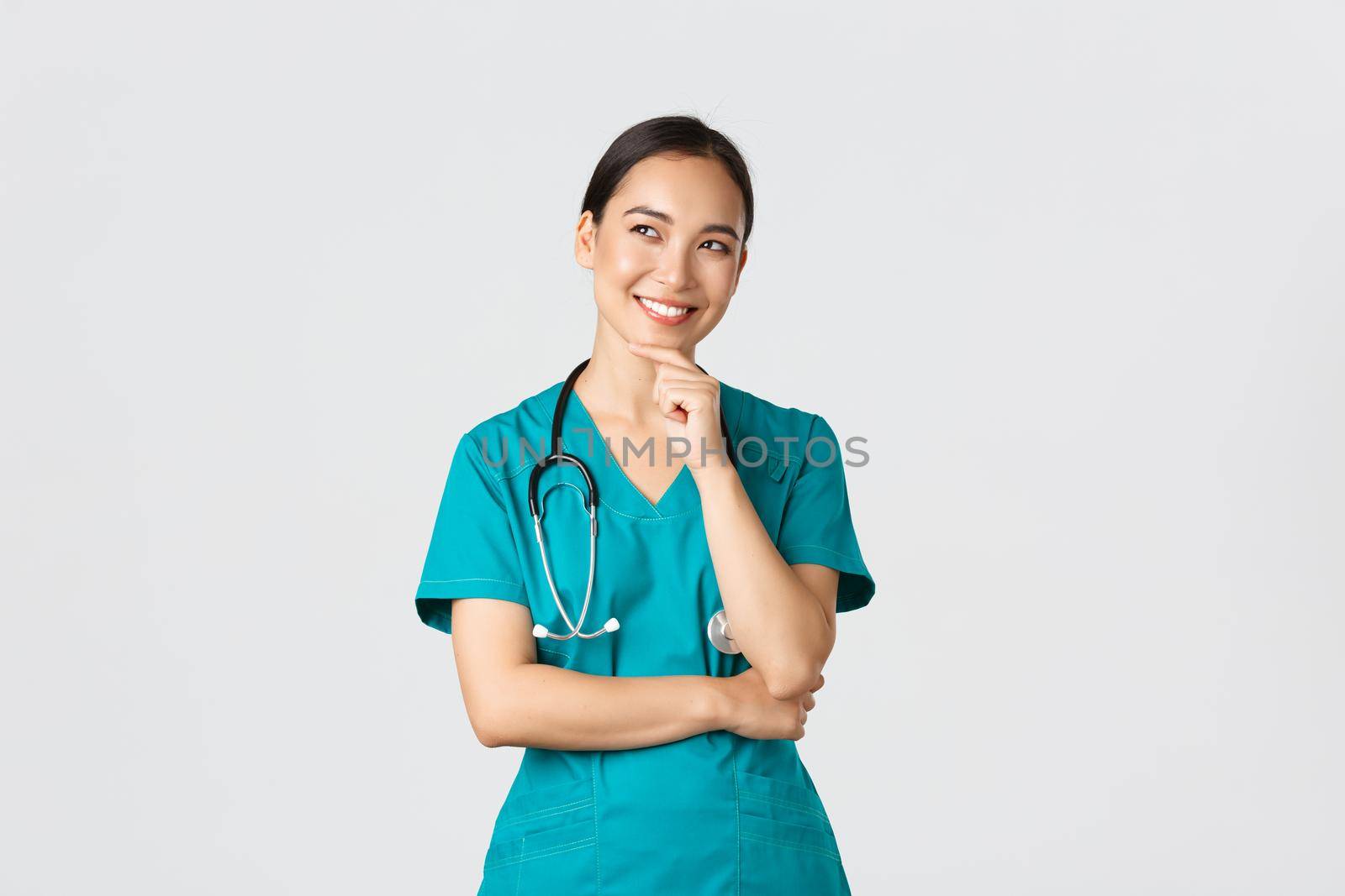 Covid-19, healthcare workers, pandemic concept. Smiling pleased attractive asian female doctor in scrubs, looking upper left corner and thinking, have idea, standing thoughtful white background.