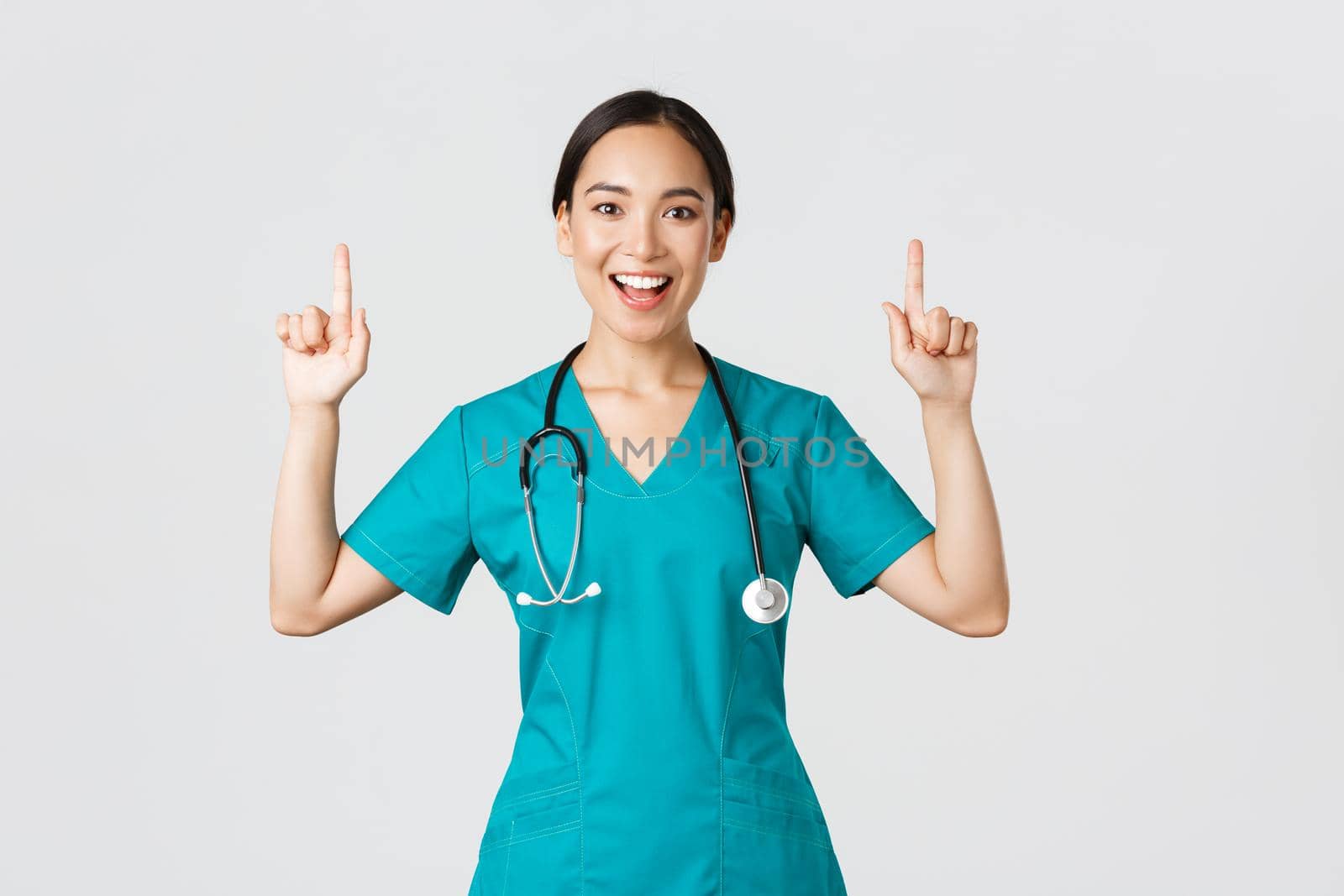 Covid-19, healthcare workers, pandemic concept. Smiling cheerful pretty nurse, female doctor or intern in scrubs pointing fingers up, showing banner, make announcement, demonstrate advertisement.