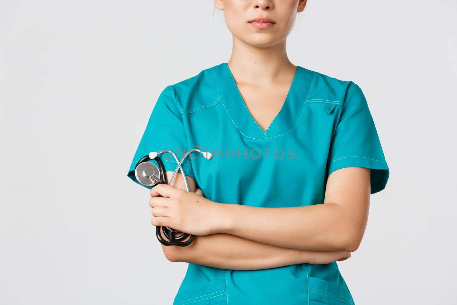 Covid-19, coronavirus disease, healthcare workers concept. Close-up of confident female asian physician, doctor in medical scrubs, cross arms determined and holding stethoscope, white background.