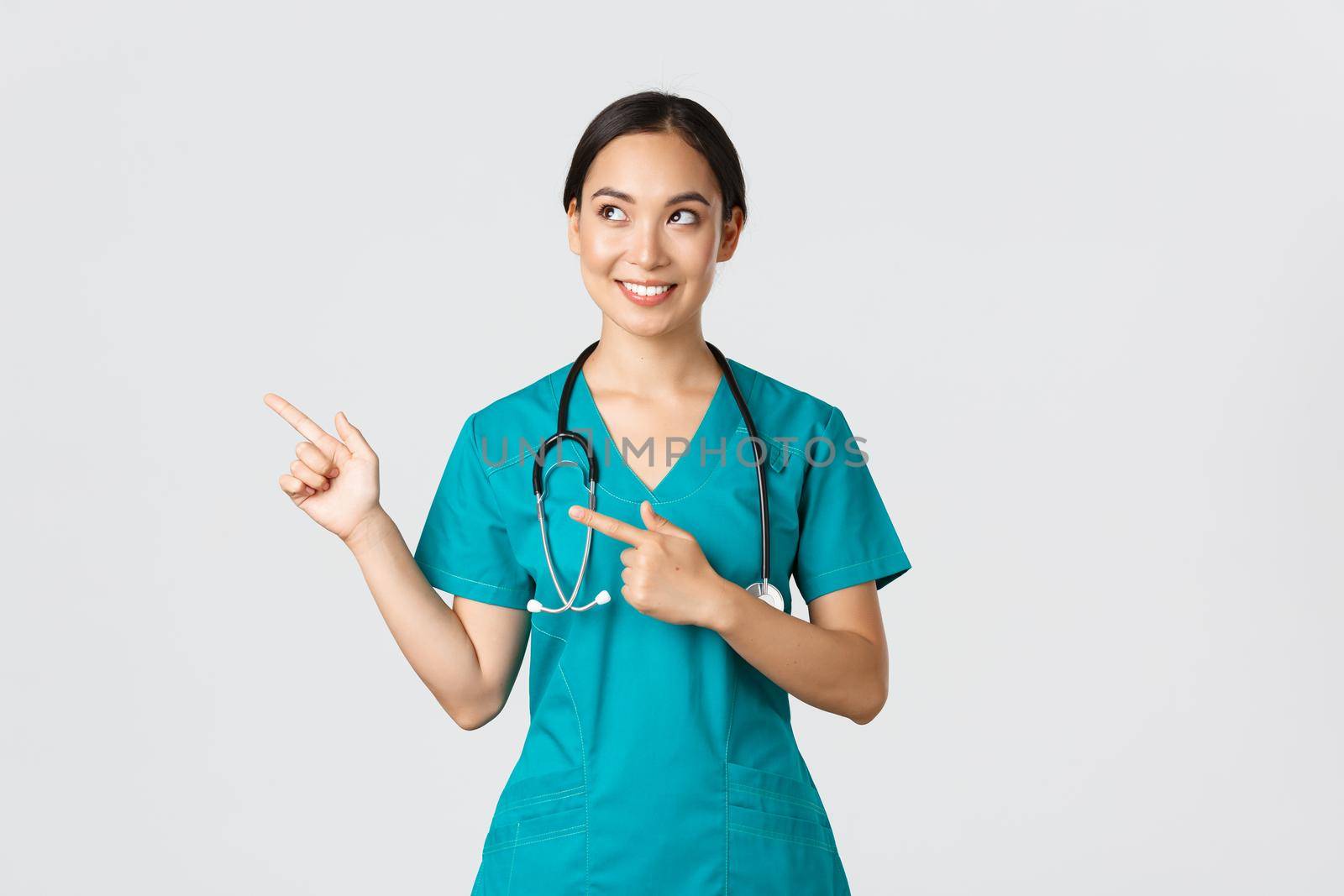 Covid-19, healthcare workers, pandemic concept. Intrigued beautiful asian female intern, nurse in scrubs looking and pointing upper left corner with curious dreamy smile, white background.
