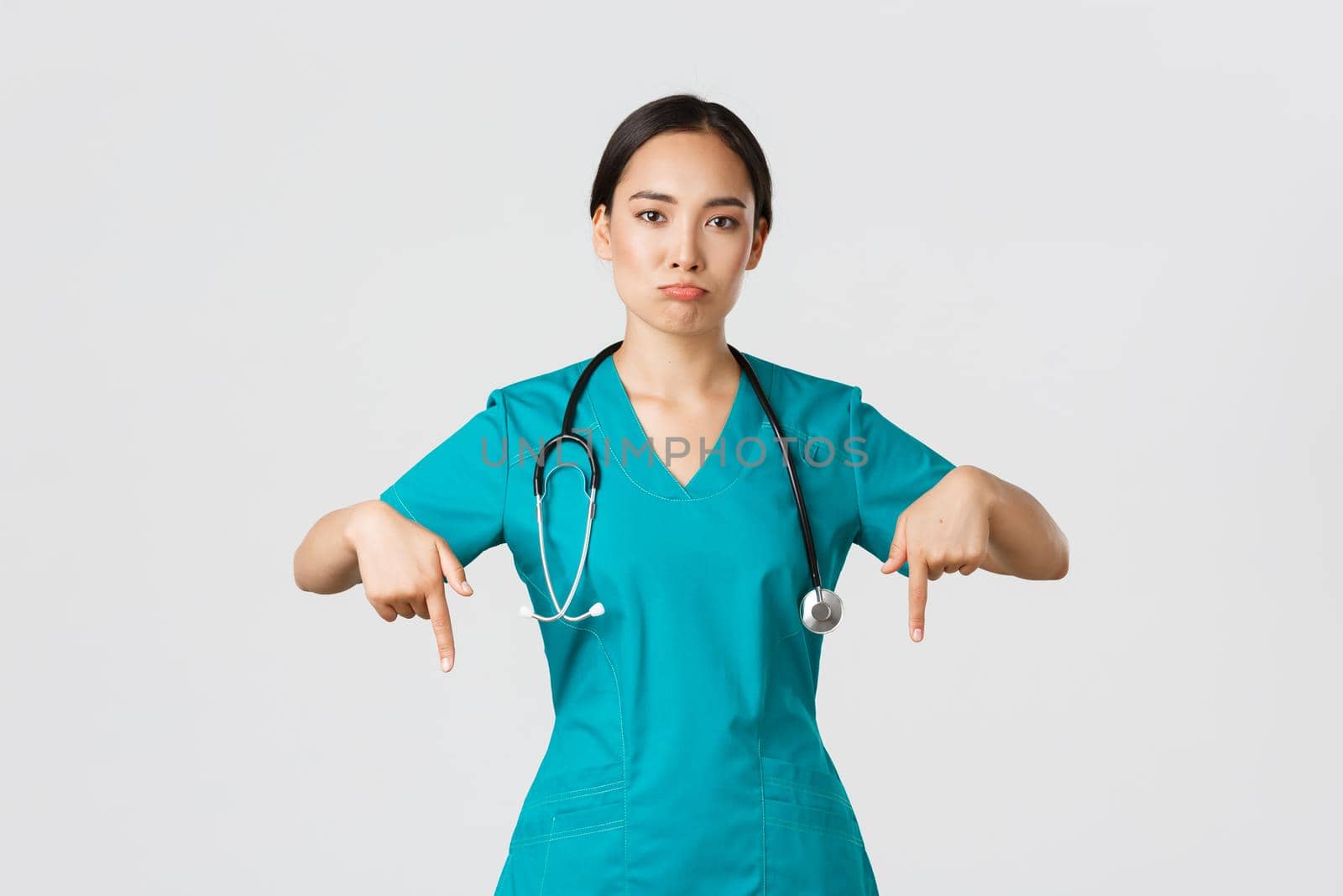 Covid-19, healthcare workers, pandemic concept. Upset and disappointed pouting asian female therapist, nurse in scrubs pointing fingers down, sulking sad, showing something with regret.