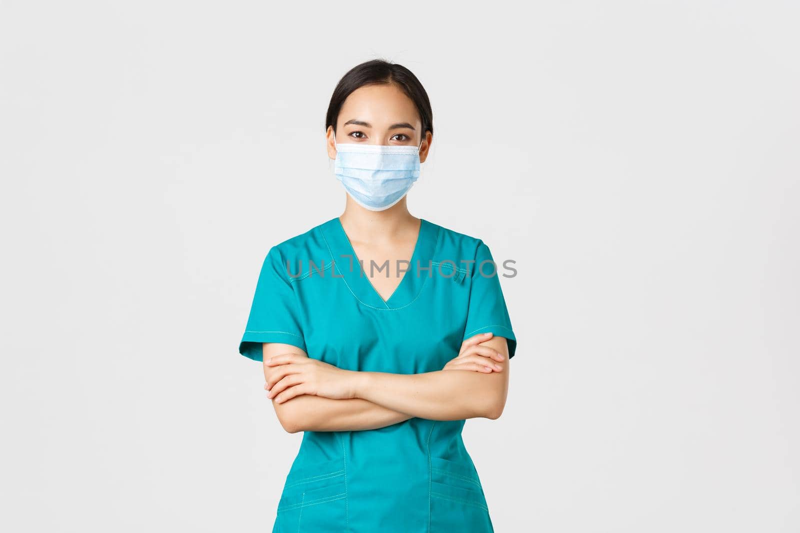 Covid-19, coronavirus disease, healthcare workers concept. Smiling confident asian female doctor, physician making checkup, wearing scrubs and medical mask, looking determined, white background.