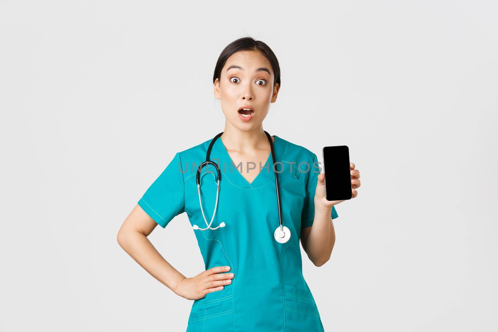 Covid-19, healthcare workers and online medicine concept. Excited and amazed asian female nurse, doctor look surprised while showing mobile phone screen, internet consultation app, white background by Benzoix
