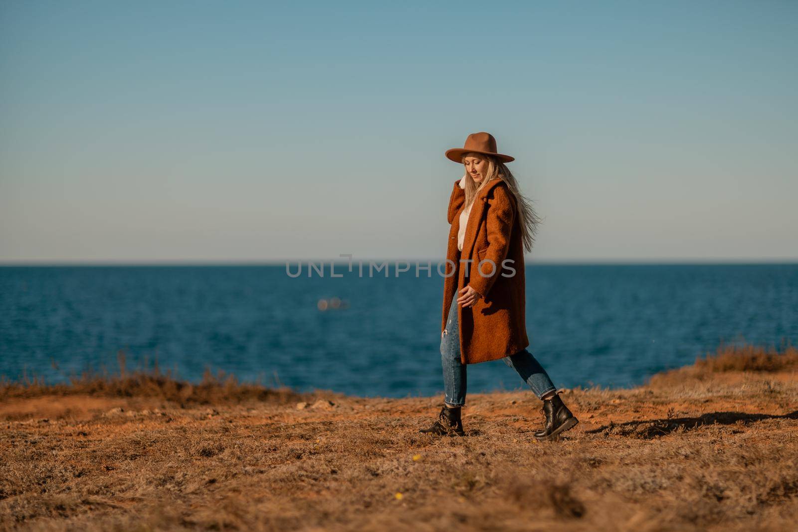 A woman walking along the coast near the sea. An elegant lady in a brown coat and a hat with fashionable makeup walks on the seashore.