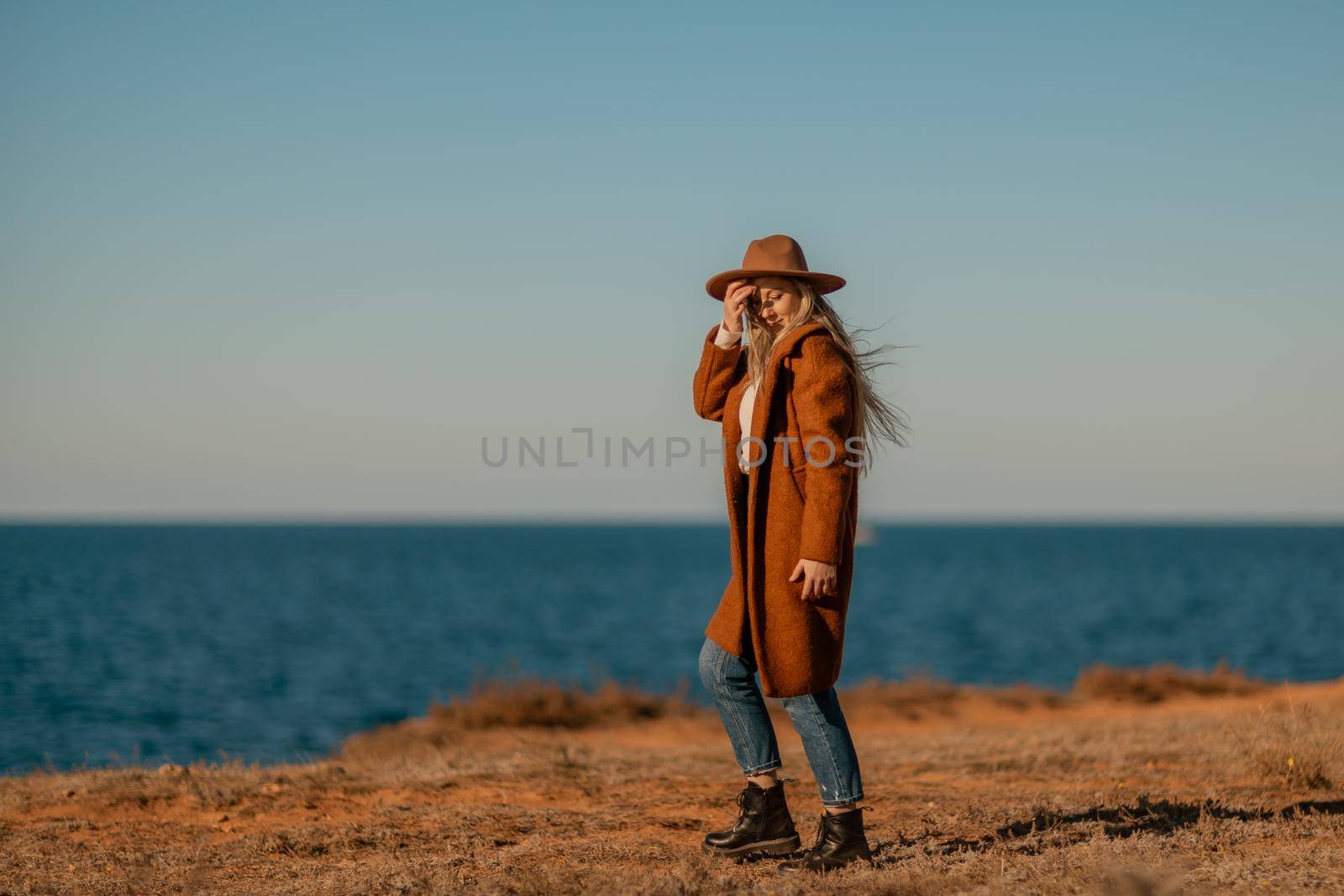 A woman walking along the coast near the sea. An elegant lady in a brown coat and a hat with fashionable makeup walks on the seashore.