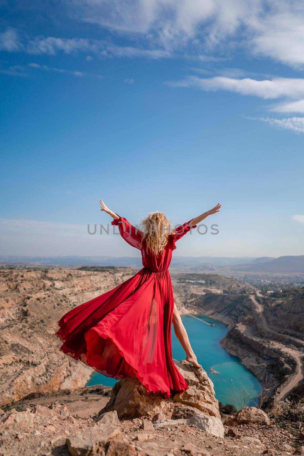 Rear view of a beautiful sensual woman in a red long dress posing on a rock high above the lake in the afternoon. Against the background of the blue sky and the lake in the shape of a heart by Matiunina