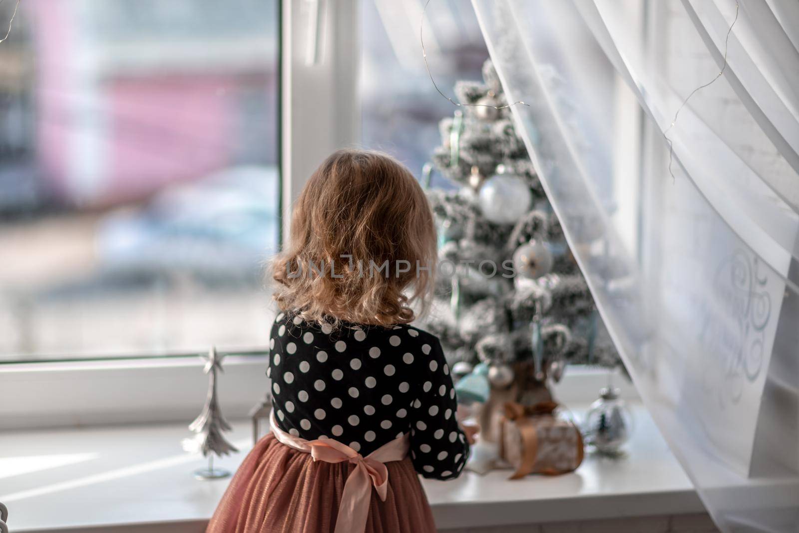 A little girl is sitting on the bed by the window and decorating a small tree with tiny Christmas toys. Happy healthy child celebrating a traditional family holiday. Adorable baby