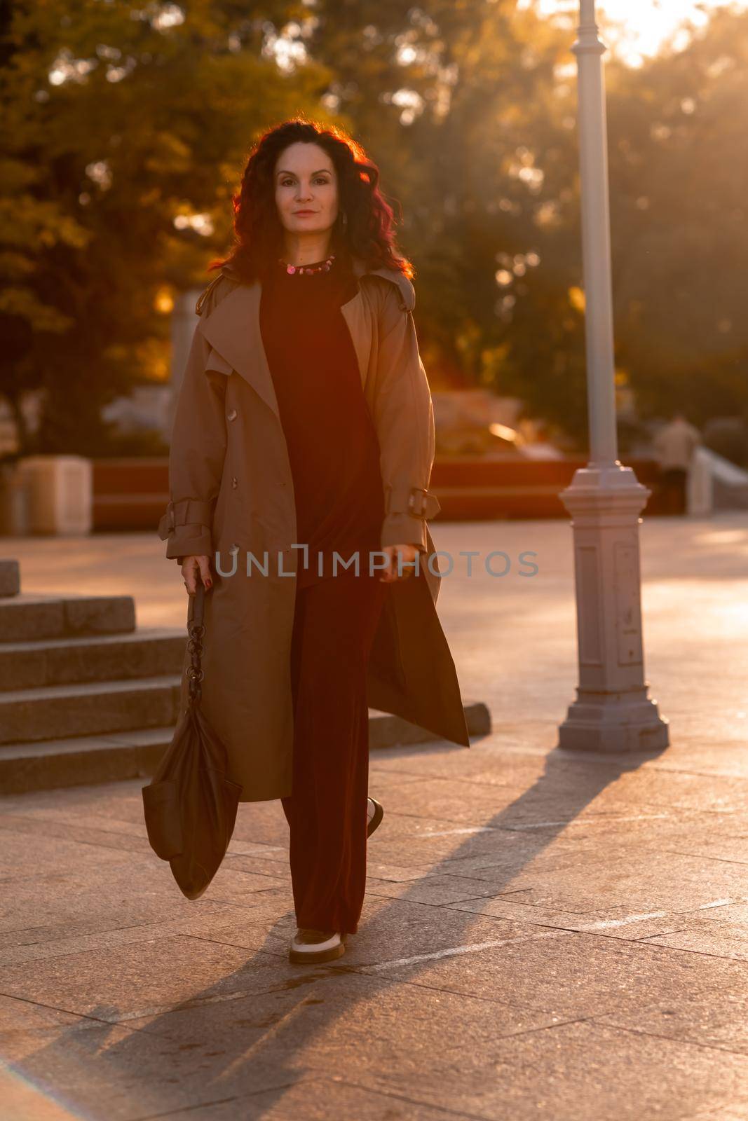 Outdoor fashion portrait of young elegant fashionable brunette woman, model in stylish hat, choker and light raincoat posing at sunset in European city. by Matiunina