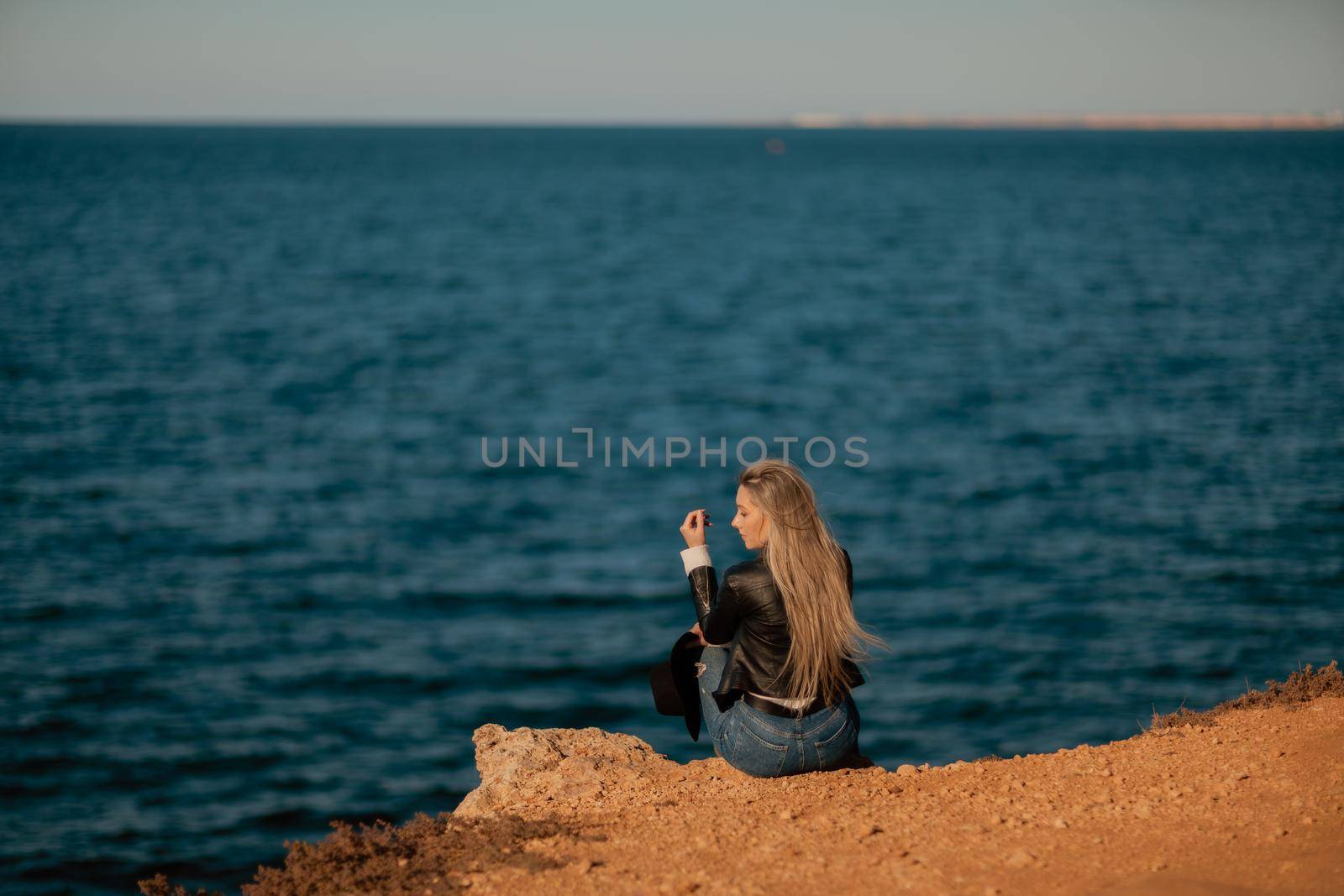 A blonde girl in a stylish black leather jacket is sitting with her back to the seashore