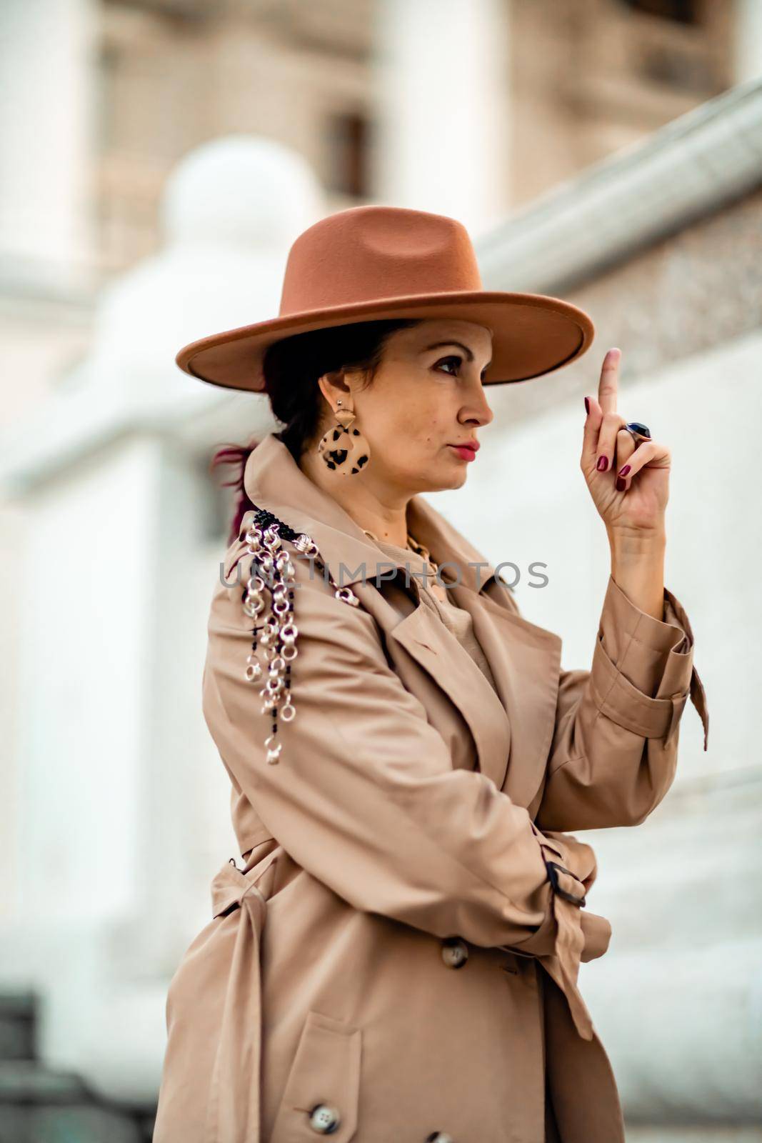 Outdoor fashion portrait of young elegant fashionable brunette woman, model in stylish hat, choker and light raincoat posing at sunset in European city