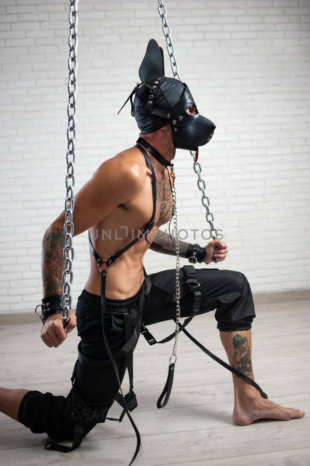 the man in a leather bdsm mask of a dog handcuffed to chains is kneeling against the wall