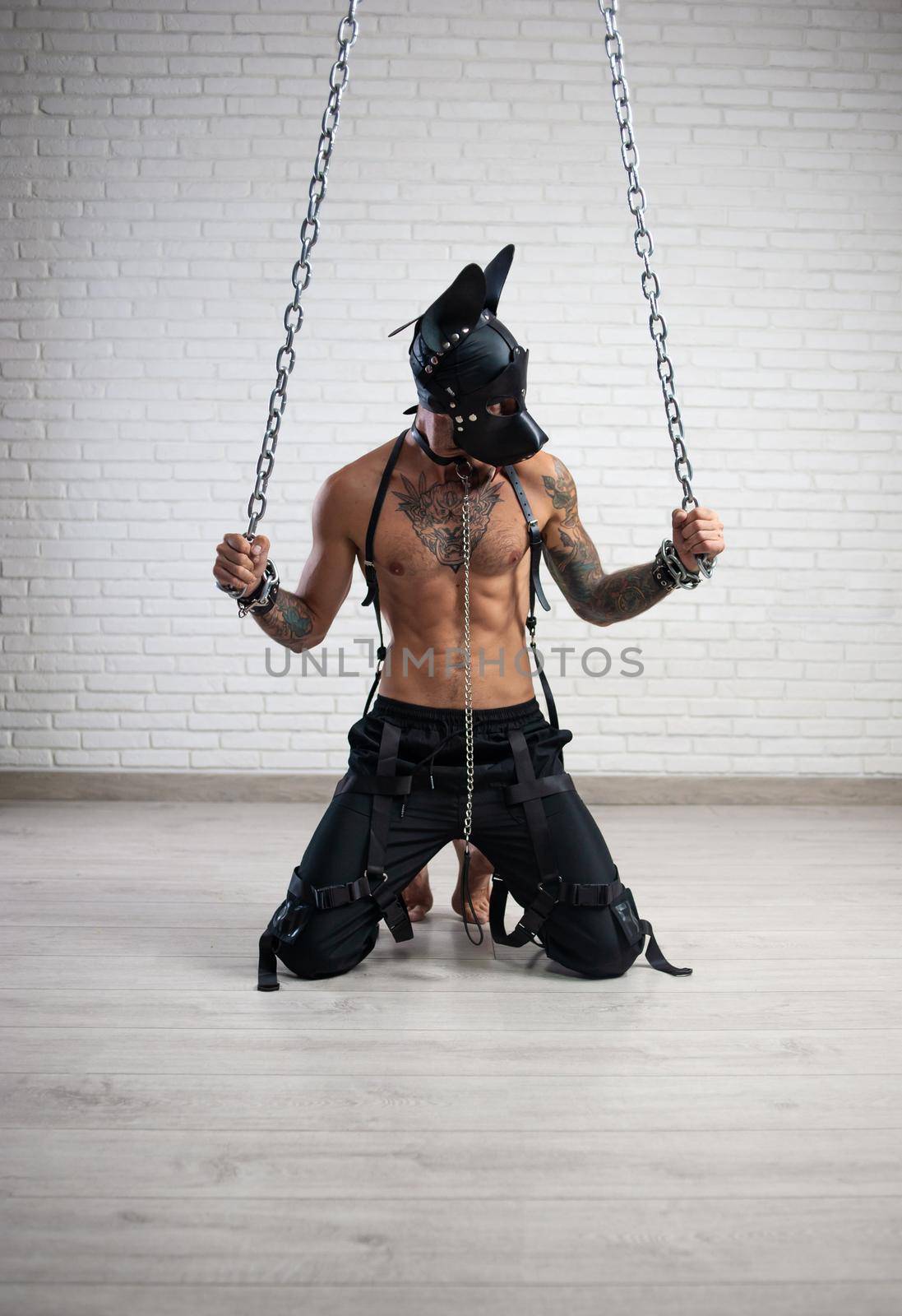 the man in a leather bdsm mask of a dog handcuffed to chains is kneeling against the wall