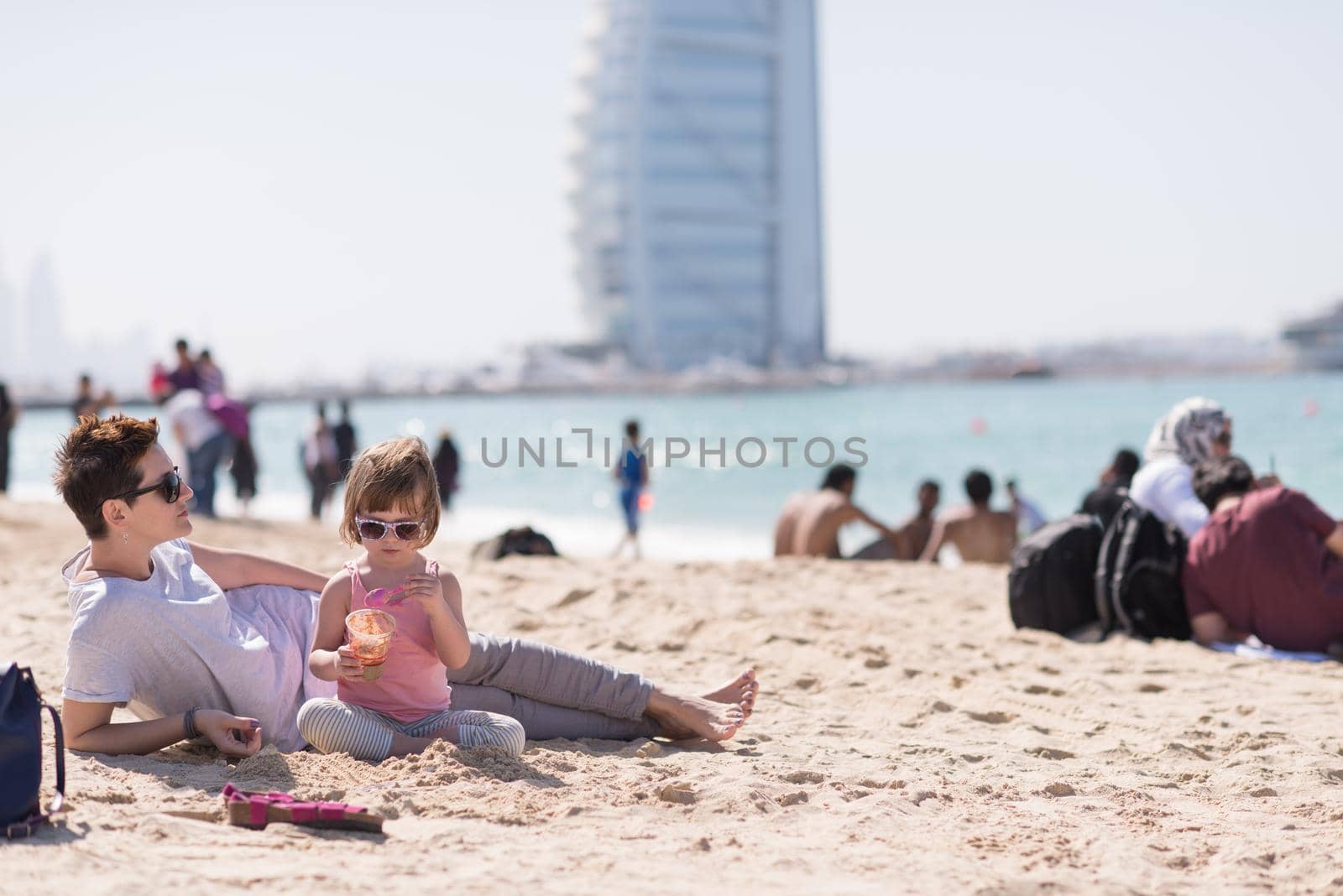 cool young mom and her little girl playing with a bucket full of sand wearing sunglasses and relaxing on the beach on a sunny day