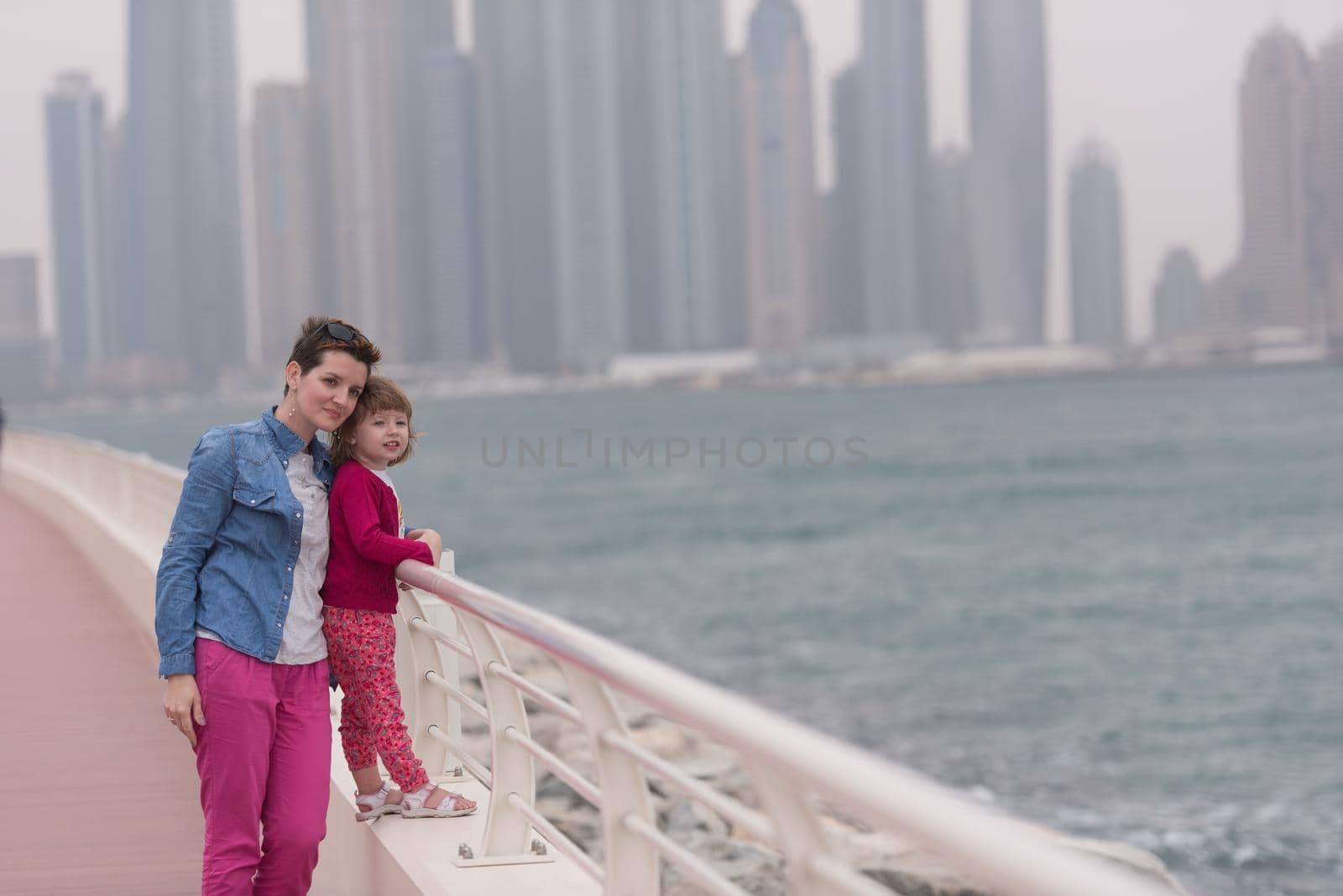 young mother and cute little girl running and cheerfully spend their time on the promenade by the sea with a big city in the background