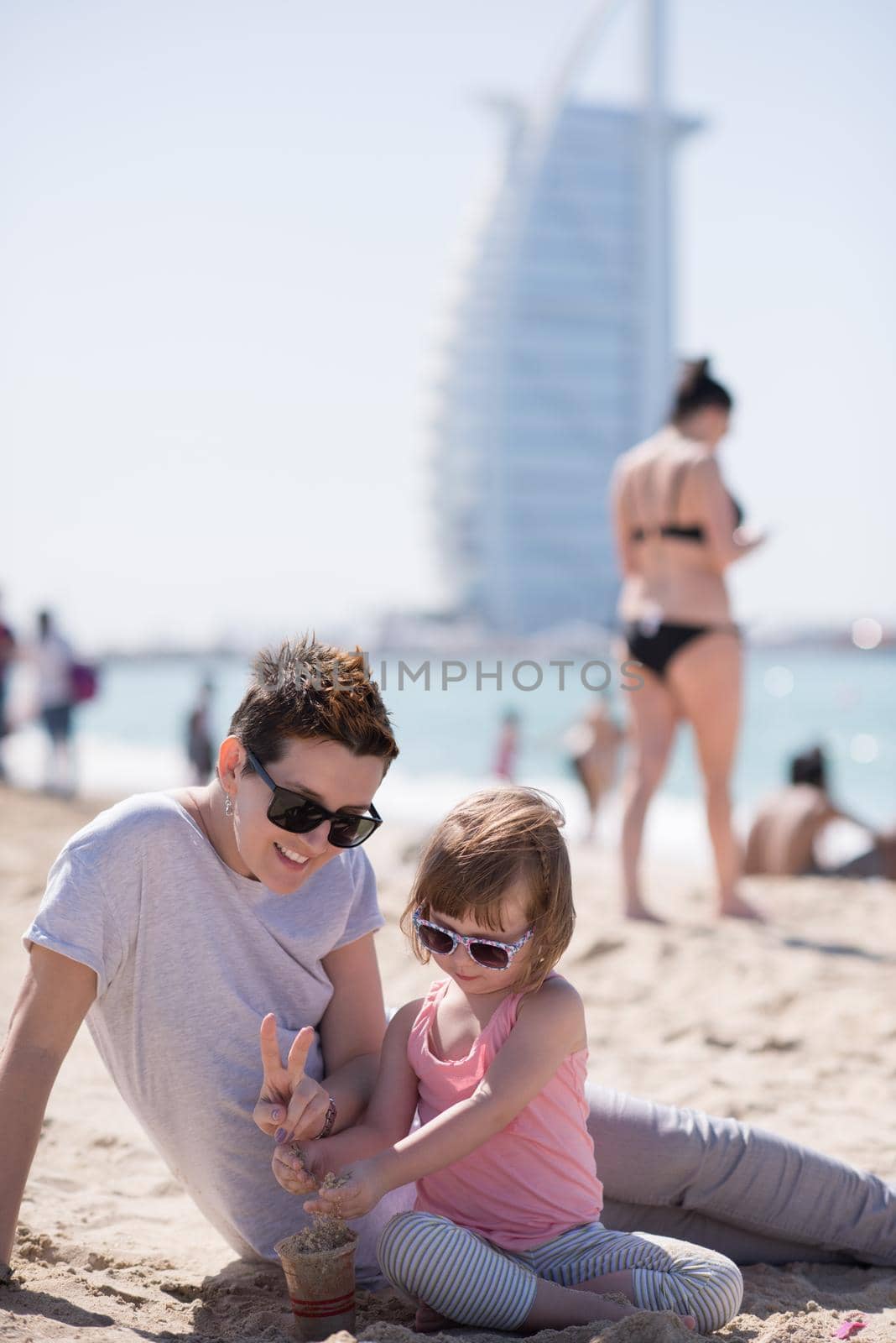 cool young mom and her little girl playing with a bucket full of sand wearing sunglasses and relaxing on the beach on a sunny day