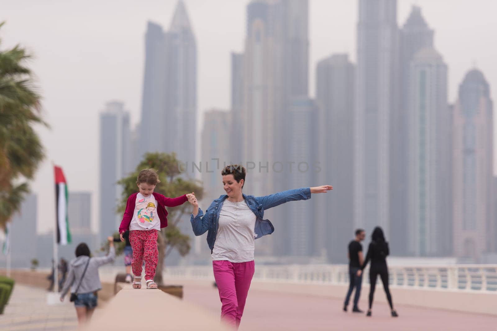 young mother and cute little girl running and cheerfully spend their time on the promenade by the sea with a big city in the background