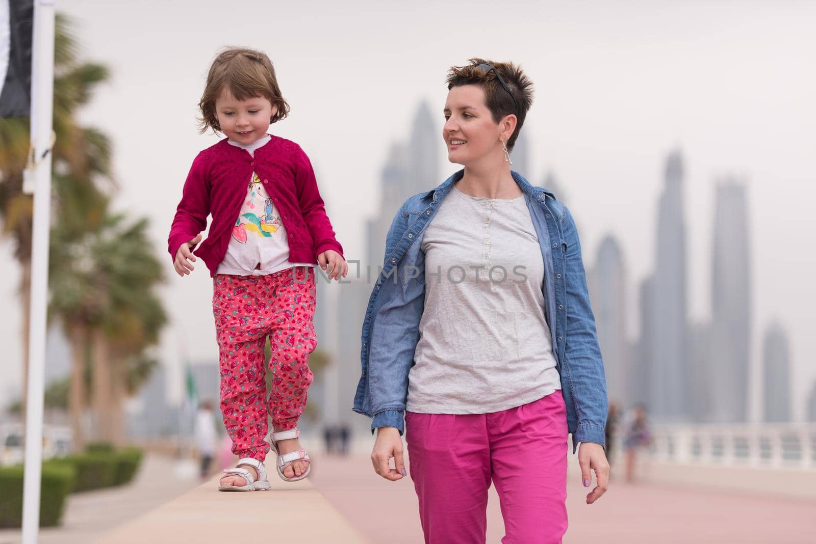 young mother and cute little girl running and cheerfully spend their time on the promenade by the sea with a big city in the background