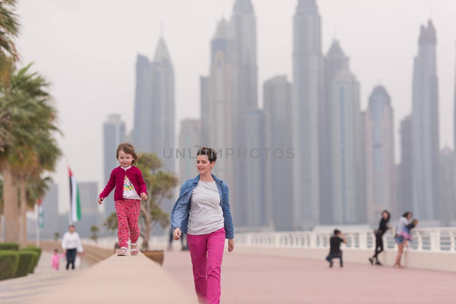 young mother and cute little girl running and cheerfully spend their time on the promenade by the sea with a big city in the background