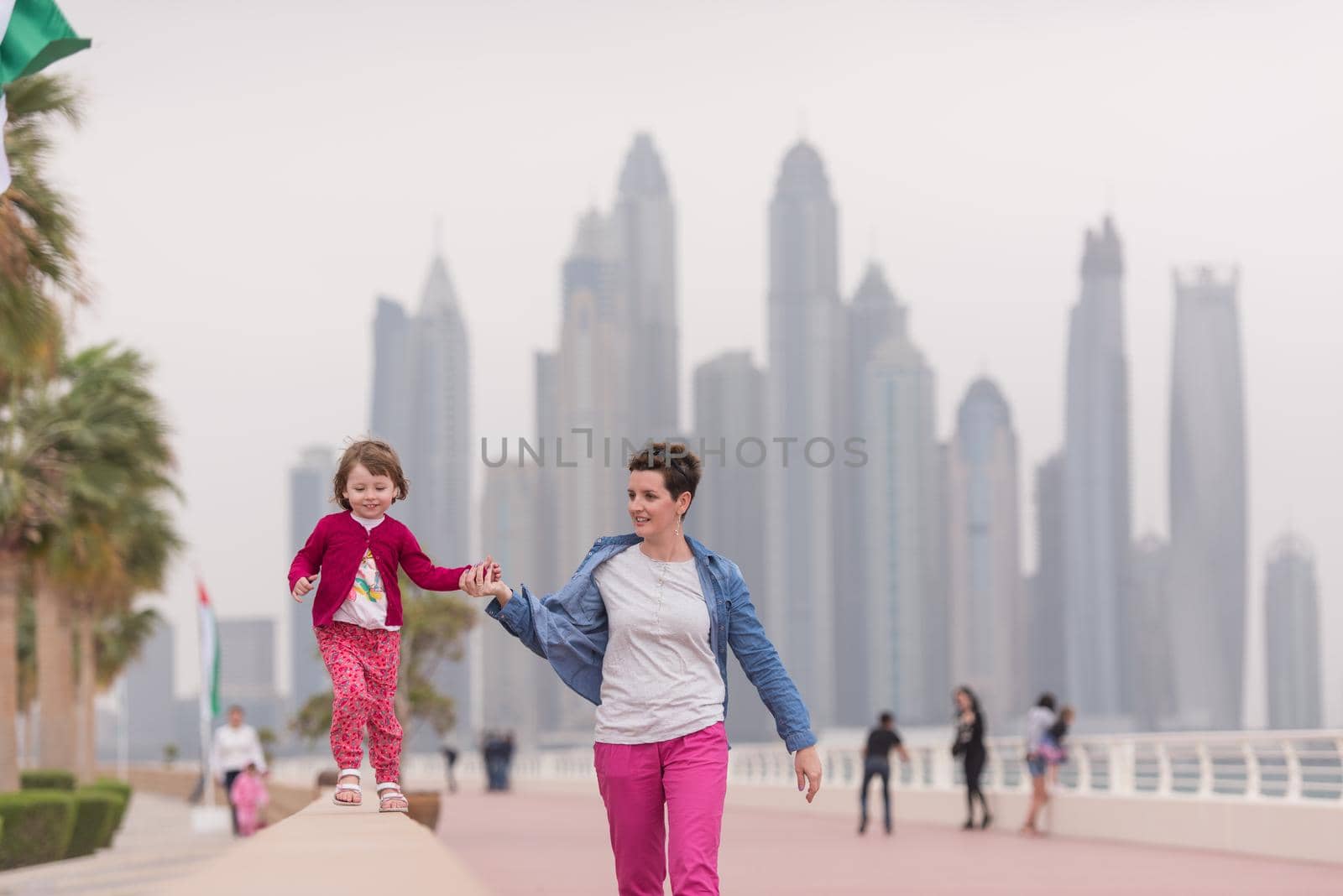 young mother and cute little girl running and cheerfully spend their time on the promenade by the sea with a big city in the background