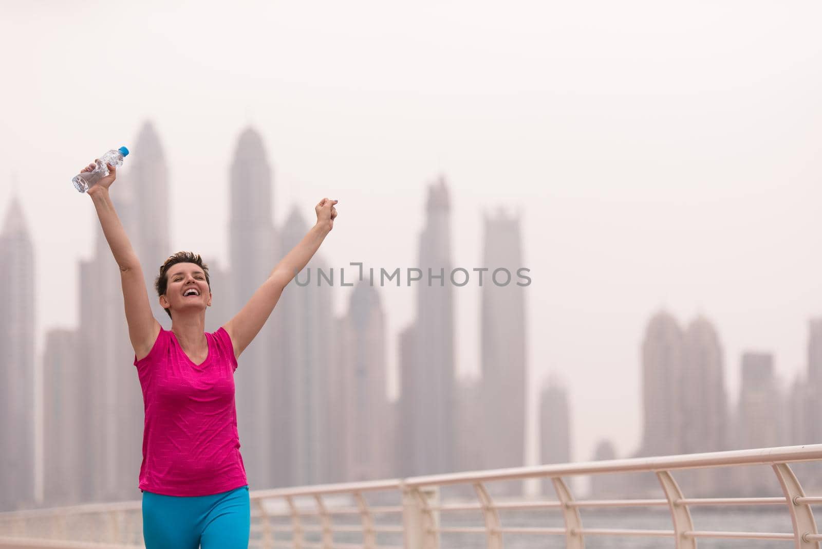 young woman celebrating a successful training run on the promenade by the sea with a bottle of water and her hands raised in the air with a big city in the background