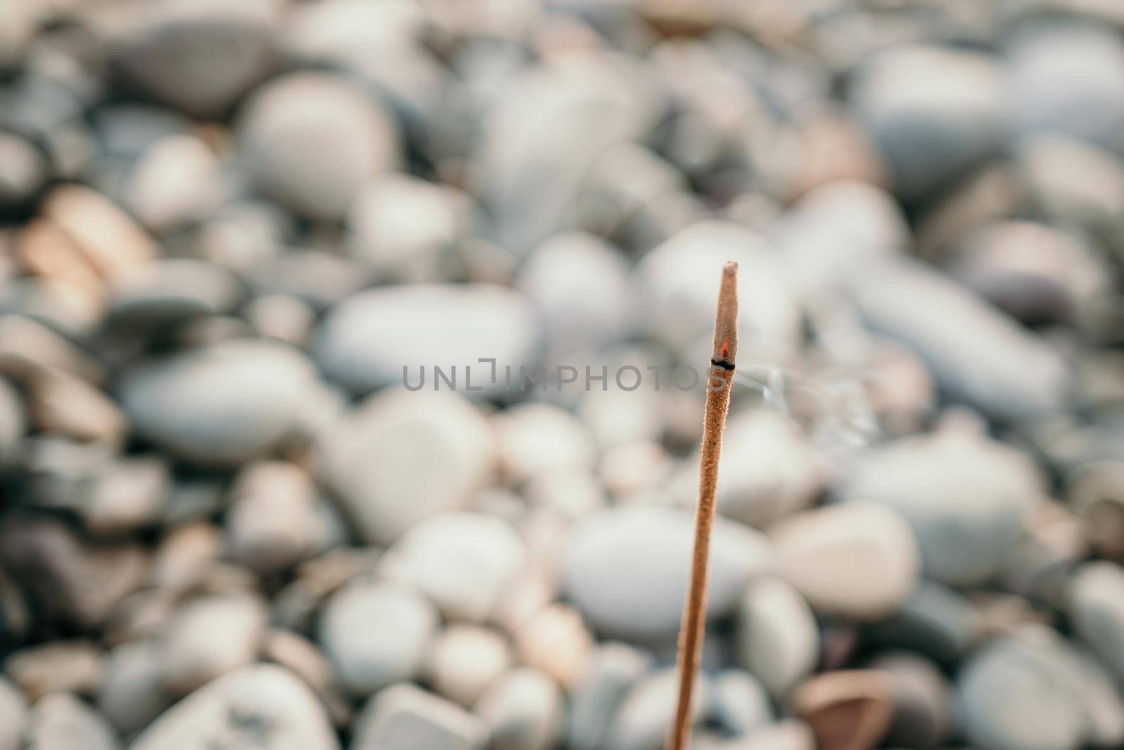 Close up of incense aroma stick burning on the pebble beach sea shore. Small waves on water surface in motion blur with bokeh lights from sunrise. Meditation concept. by panophotograph