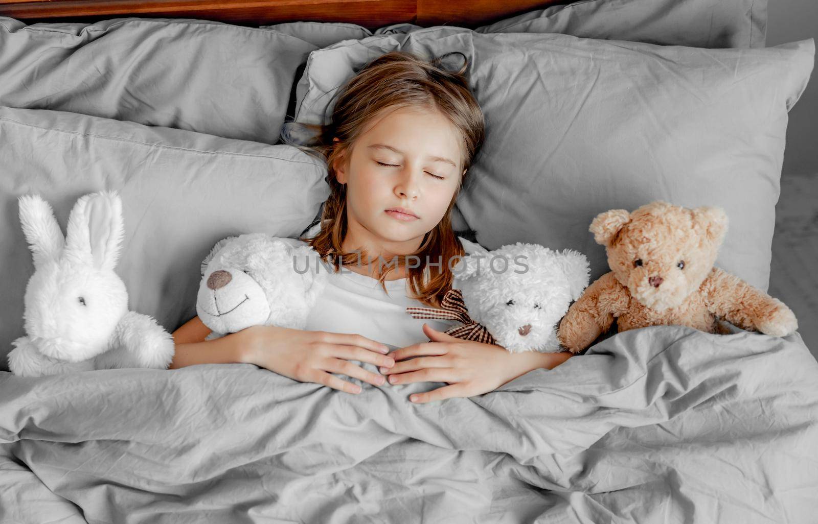 Beautiful child girl sleeping in the bed with teddy bear and toys. Pretty kid napping in bedchamber under blanket