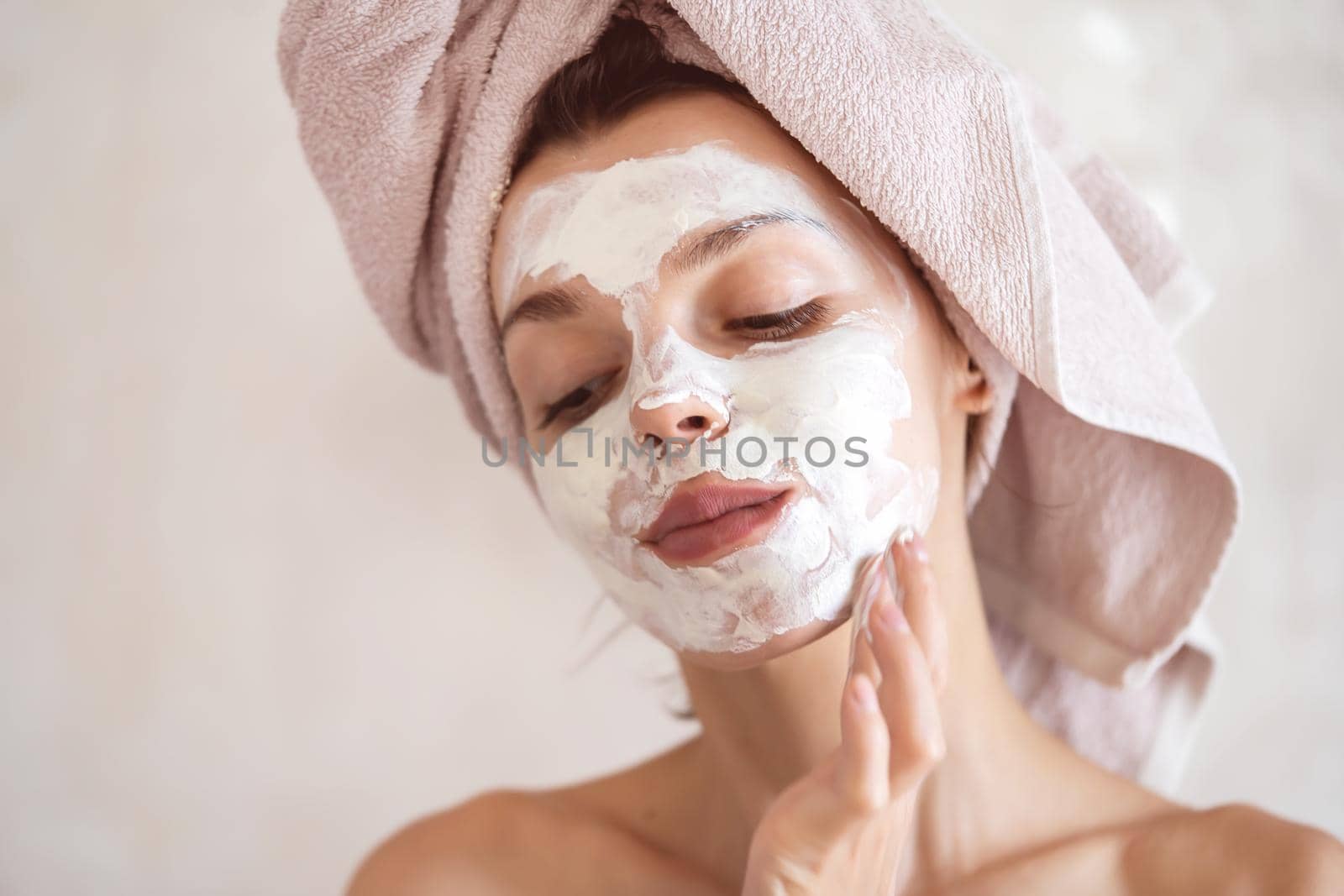 A young girl in a towel on her head and body stands near the mirror in the bathroom and applies a clay mask to her face, the woman takes care of her health and beauty.