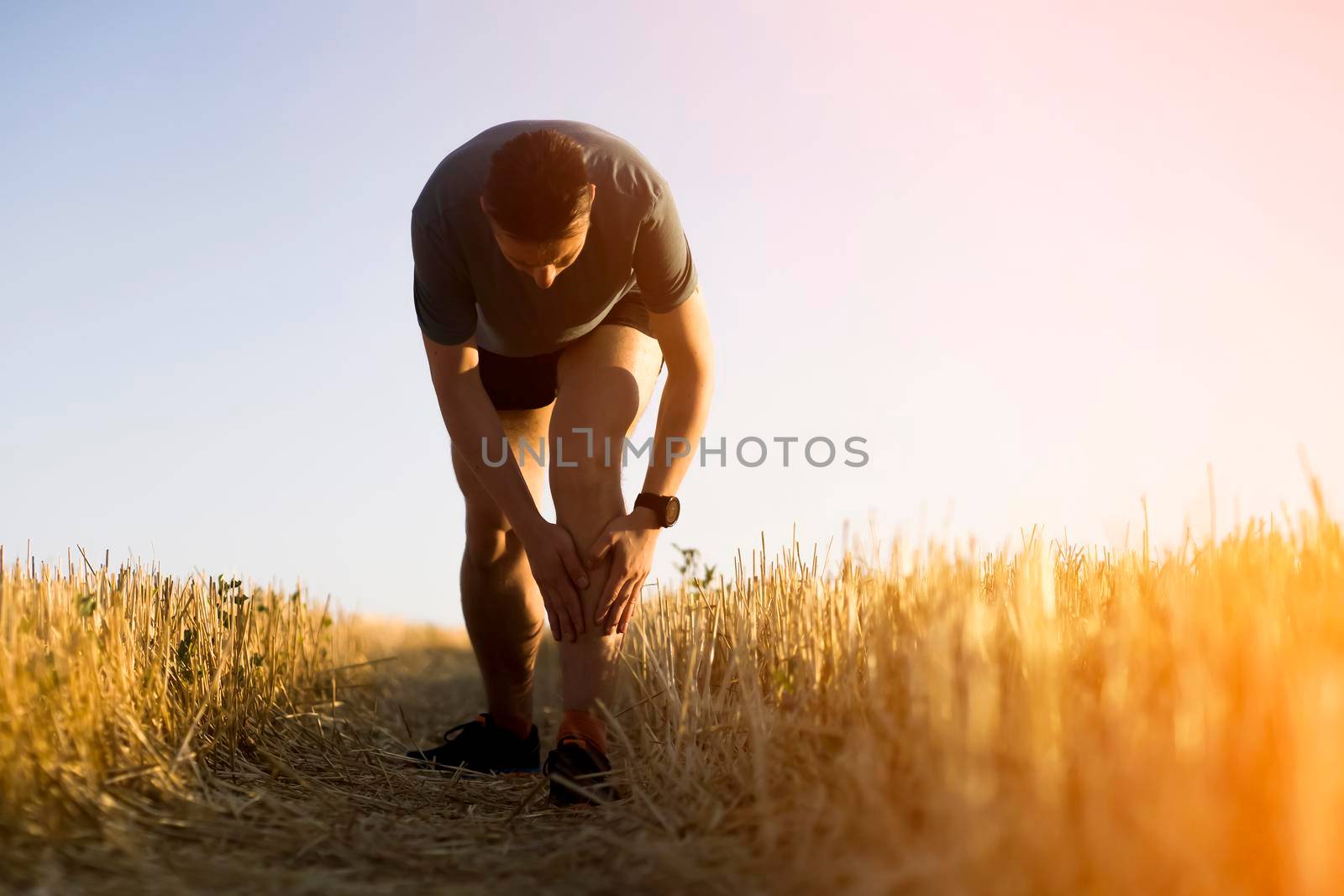 A young man jogging at sunset on the trail across the field, the tired athlete holds on to his sore knee.