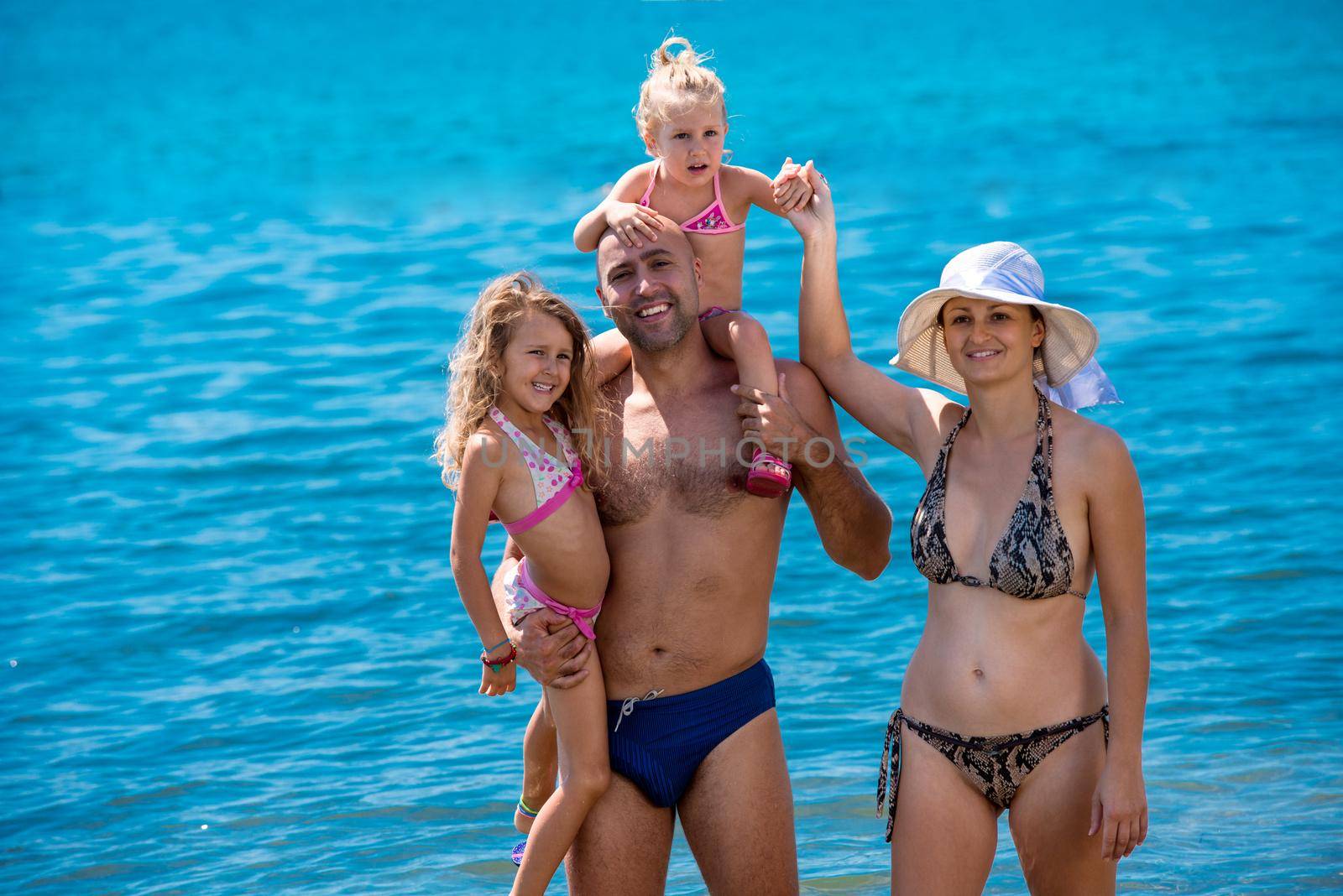 portrait of a young happy couple with cute little daughters having fun while standing in the shallow water of the sea during Summer vacation  Healthy family holiday concept