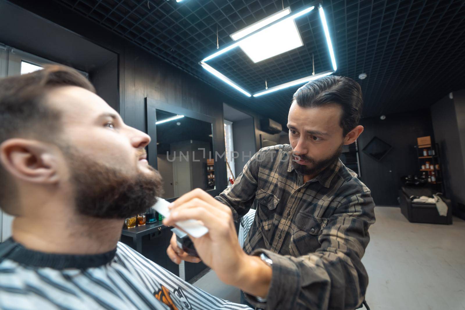 Barber works with a beard clipper. Hipster client getting haircut. by teksomolika