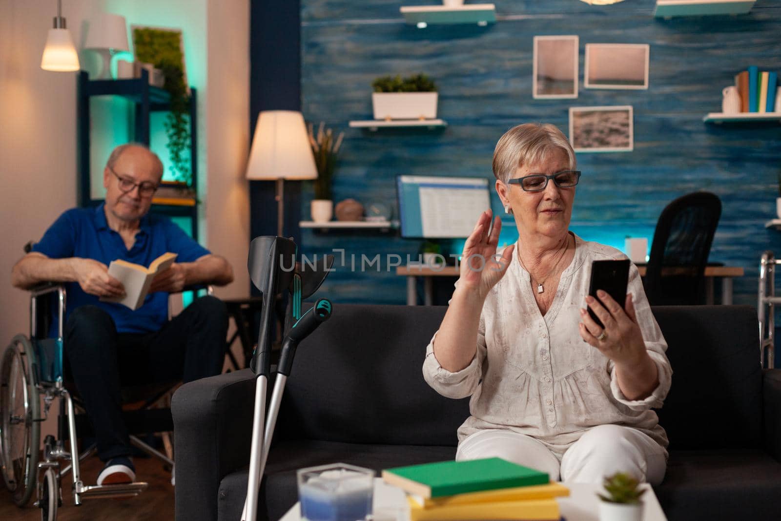 Caucasian old woman talking on video call with smartphone on living room sofa. Person using online remote conference while disabled senior man sitting in wheelchair with book in background