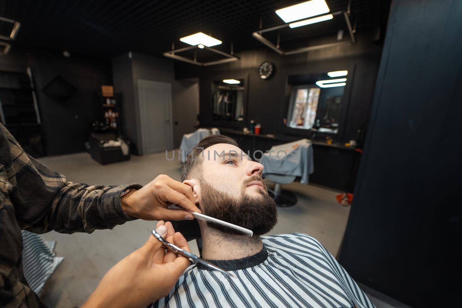 Hairdresser doing haircut of beard using comb and scissors to young attractive man in barbershop