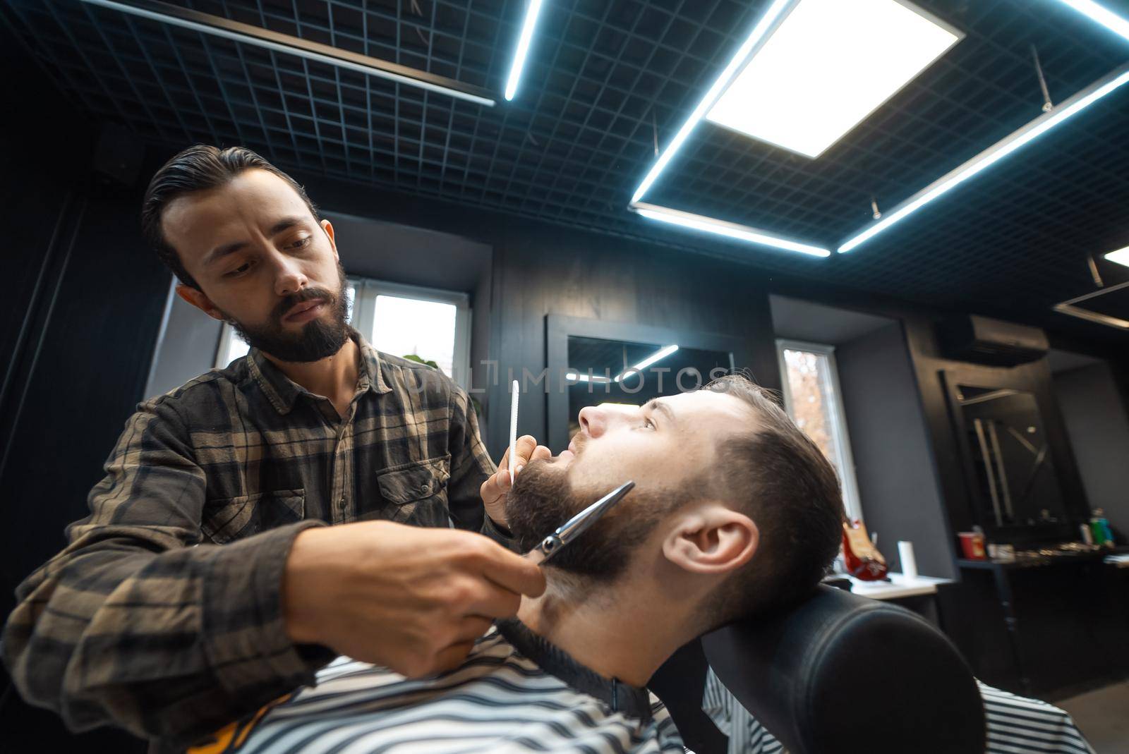 Hairdresser doing haircut of beard using comb and scissors to young attractive man in barbershop