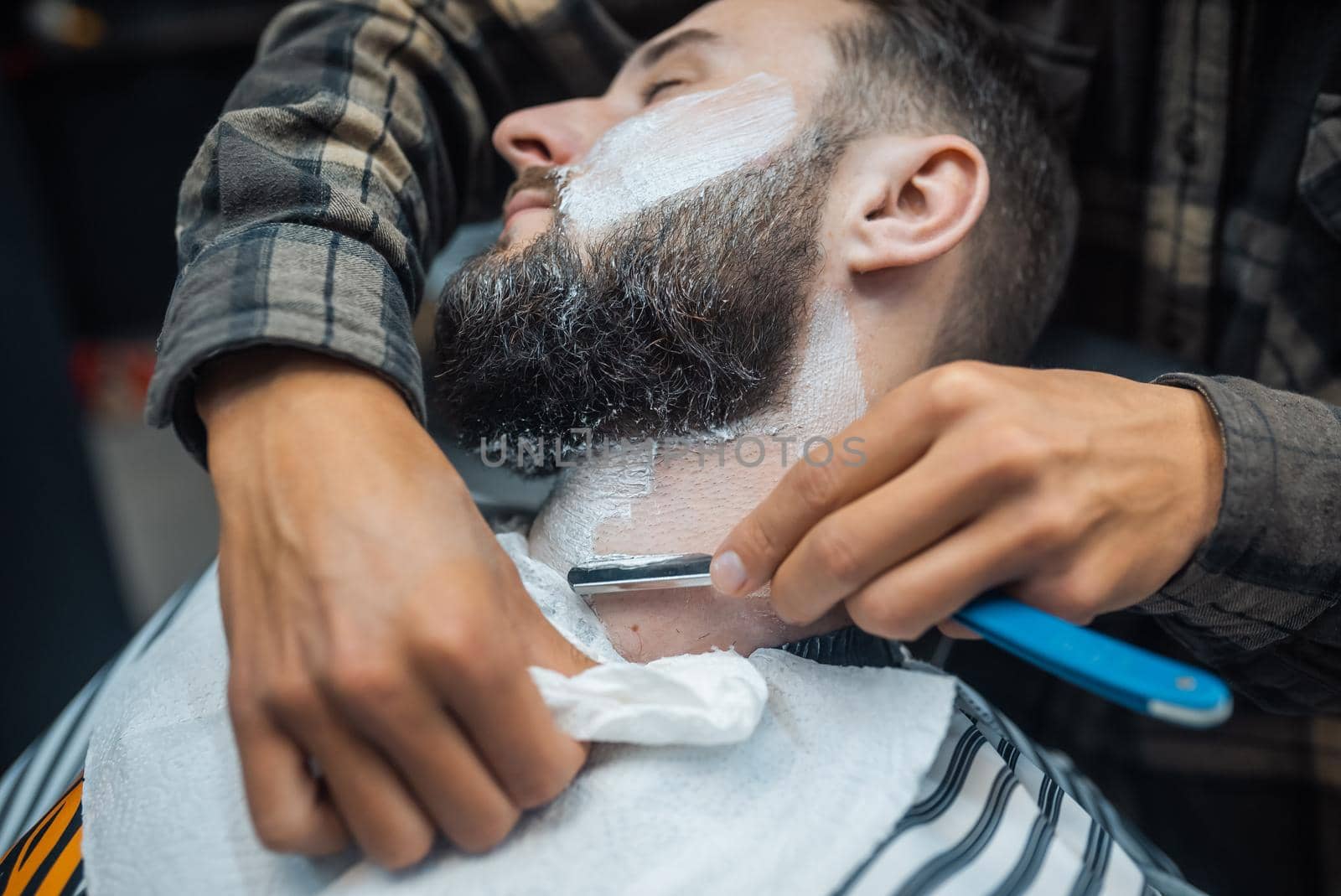 Young bearded man getting shaved by hairdresser at barbershop by teksomolika