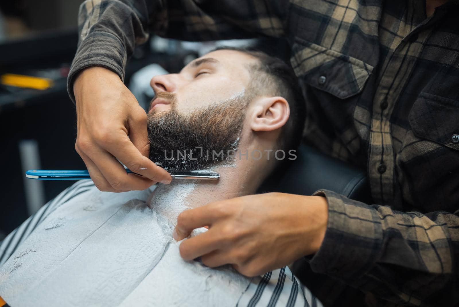 Young bearded man getting shaved with straight edge razor by hairdresser at barbershop