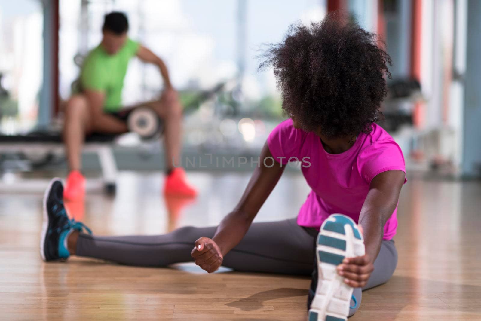 happy young african american woman in a gym stretching and warming up before workout young mab exercising with dumbbells in background