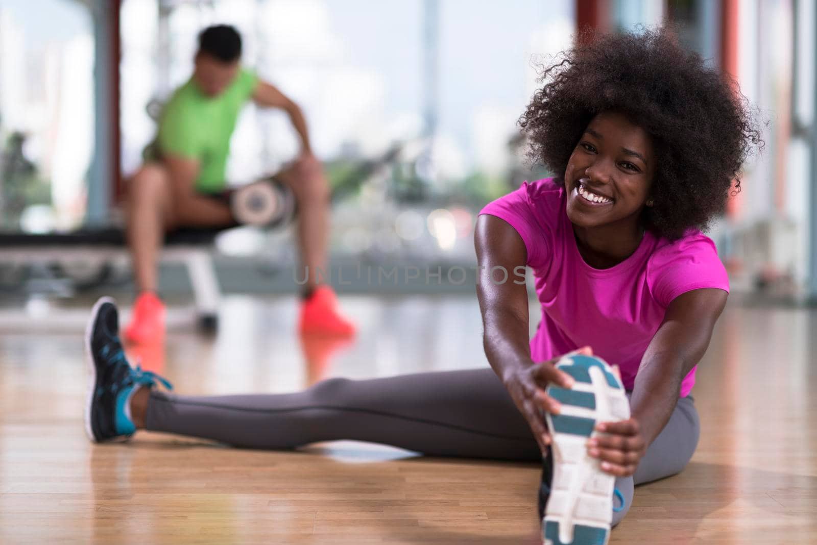 happy young african american woman in a gym stretching and warming up before workout young mab exercising with dumbbells in background