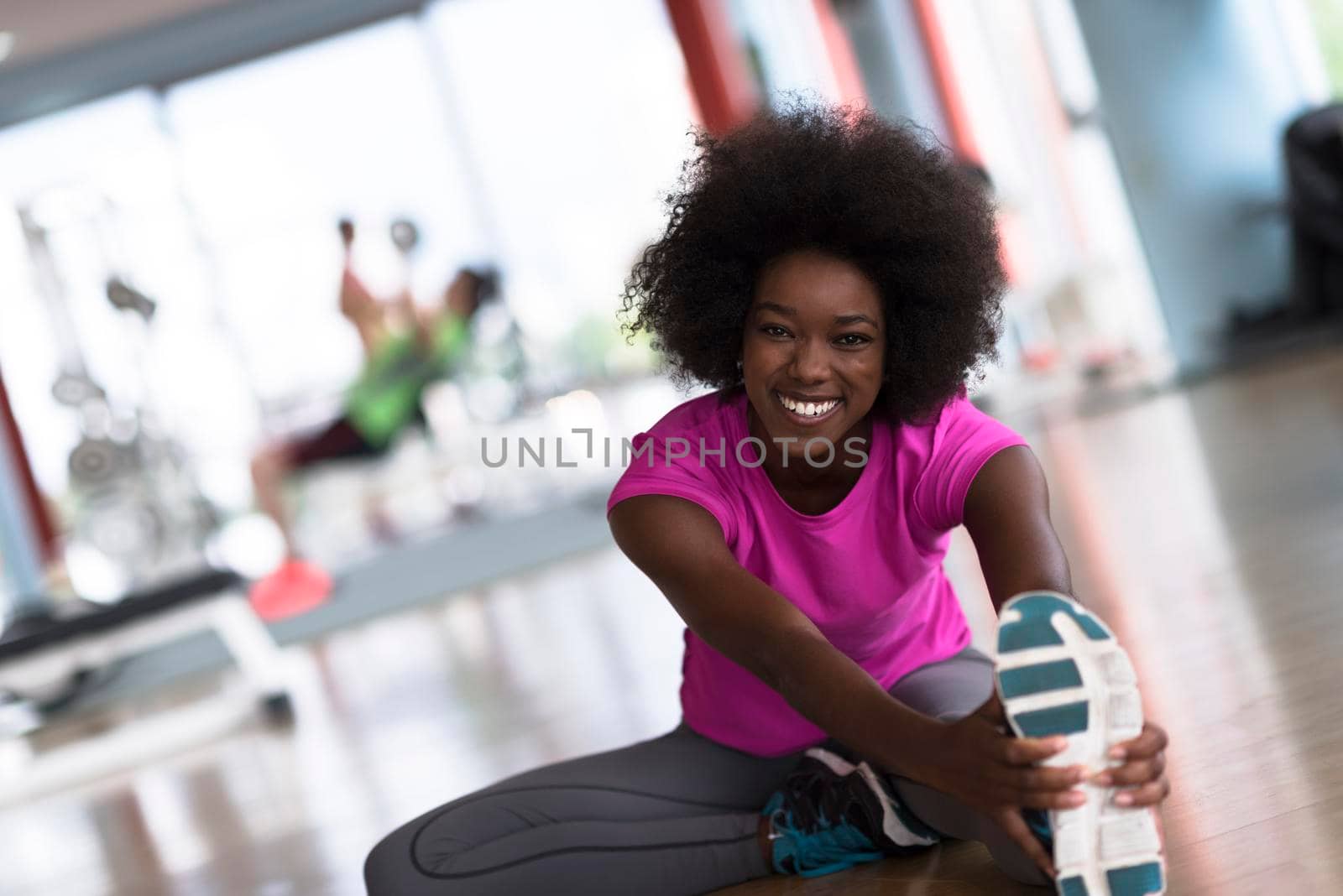 happy young african american woman in a gym stretching and warming up before workout young mab exercising with dumbbells in background