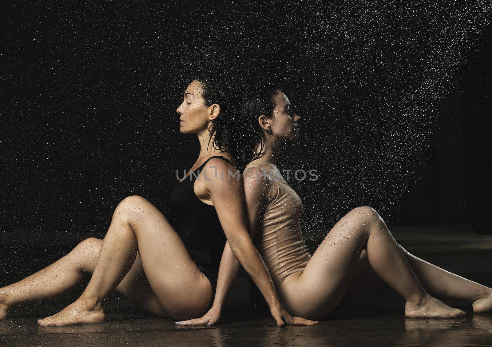 two beautiful women of Caucasian appearance with black hair sit back to back in drops of water on a black background by ndanko