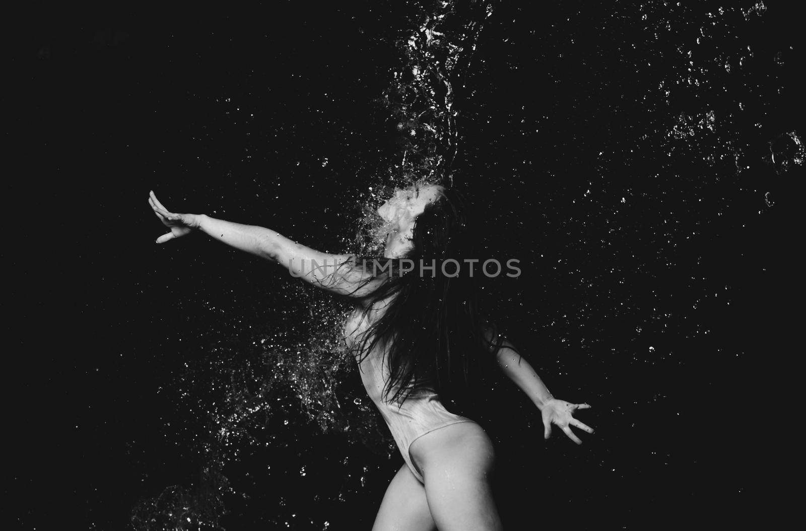 young beautiful woman of Caucasian appearance with long hair dances in drops of water on a black background. Jumping and swinging arms, dressed in a beige bodysuit