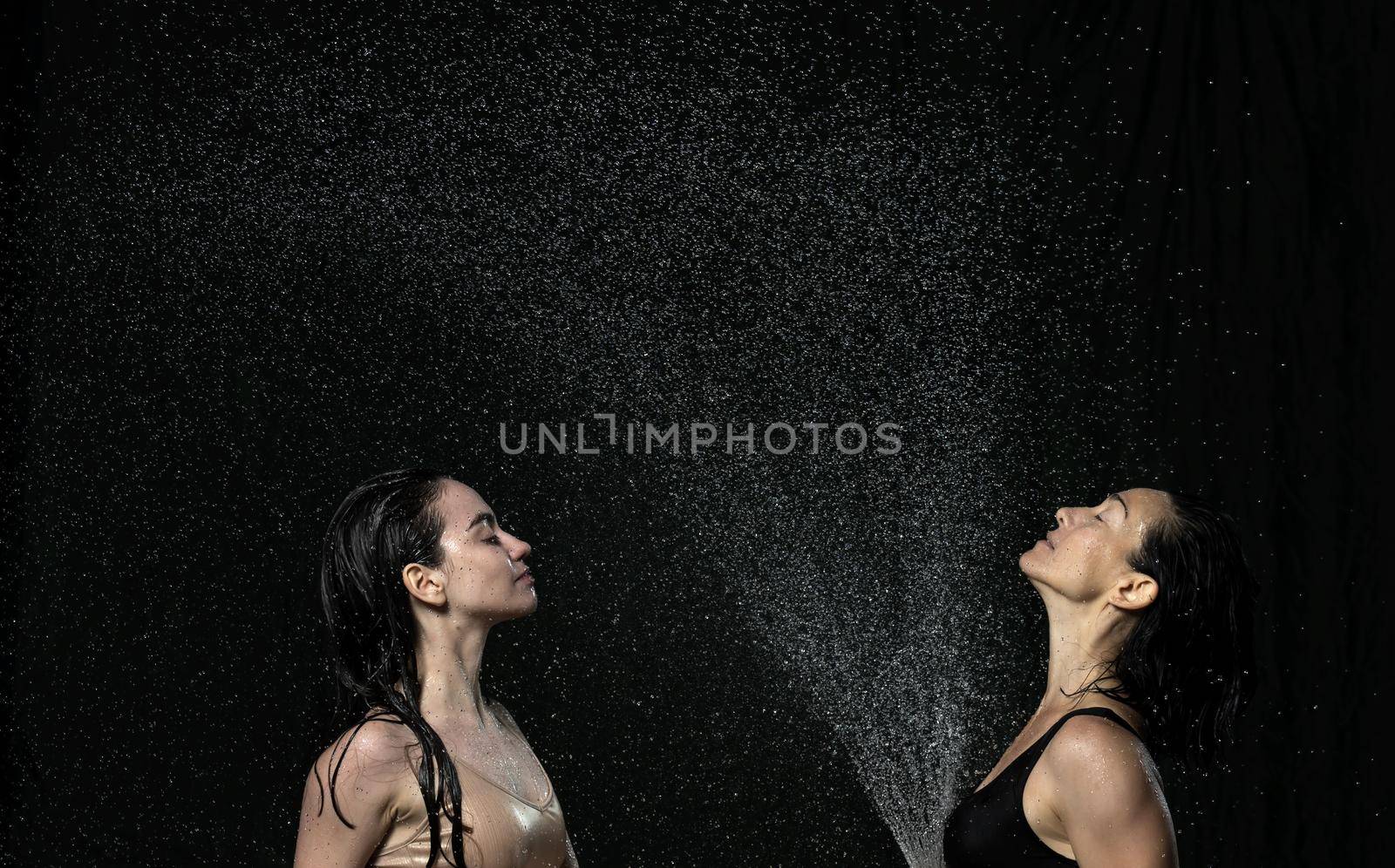 two beautiful women of Caucasian appearance with black hair sit in drops of water on a black background