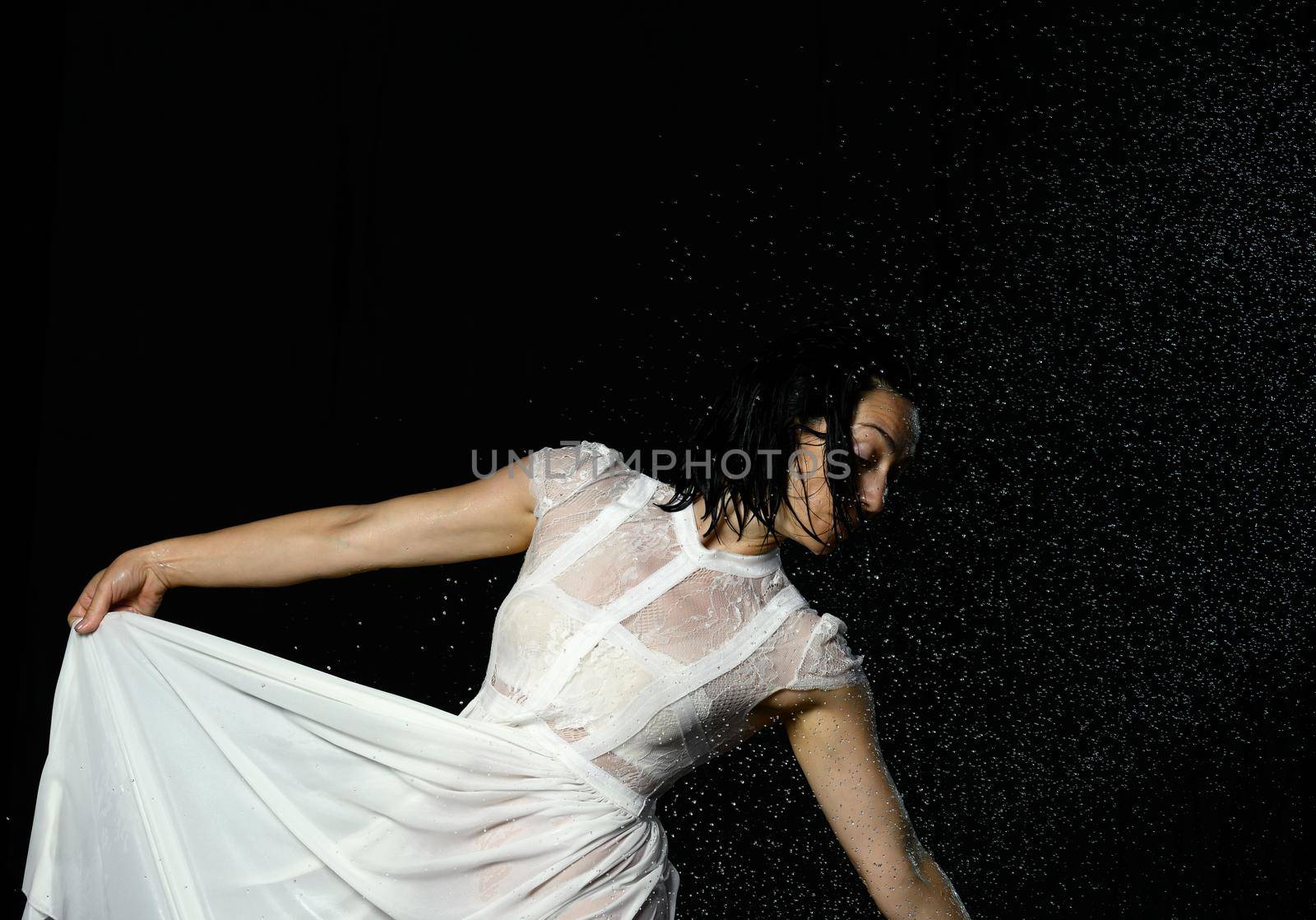 beautiful woman of Caucasian appearance with black hair dances in drops of water on a black background. The woman is wearing a white chiffon dress by ndanko
