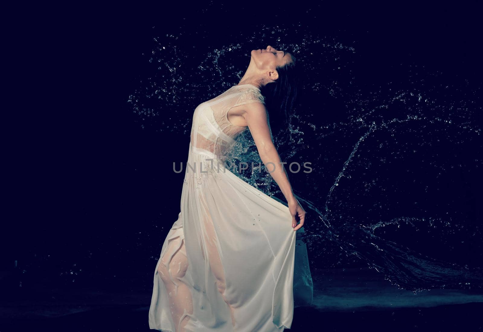 beautiful woman of Caucasian appearance with black hair dances in drops of water on a black background. The woman is wearing a white chiffon dress
