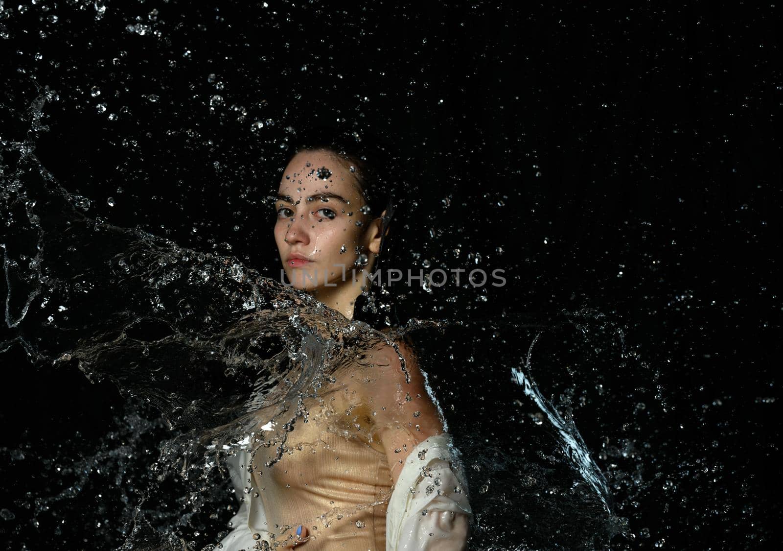 beautiful young woman of Caucasian appearance with black hair stands in drops of water on a black background. Woman wearing white shirt and looking at the camera, serious face