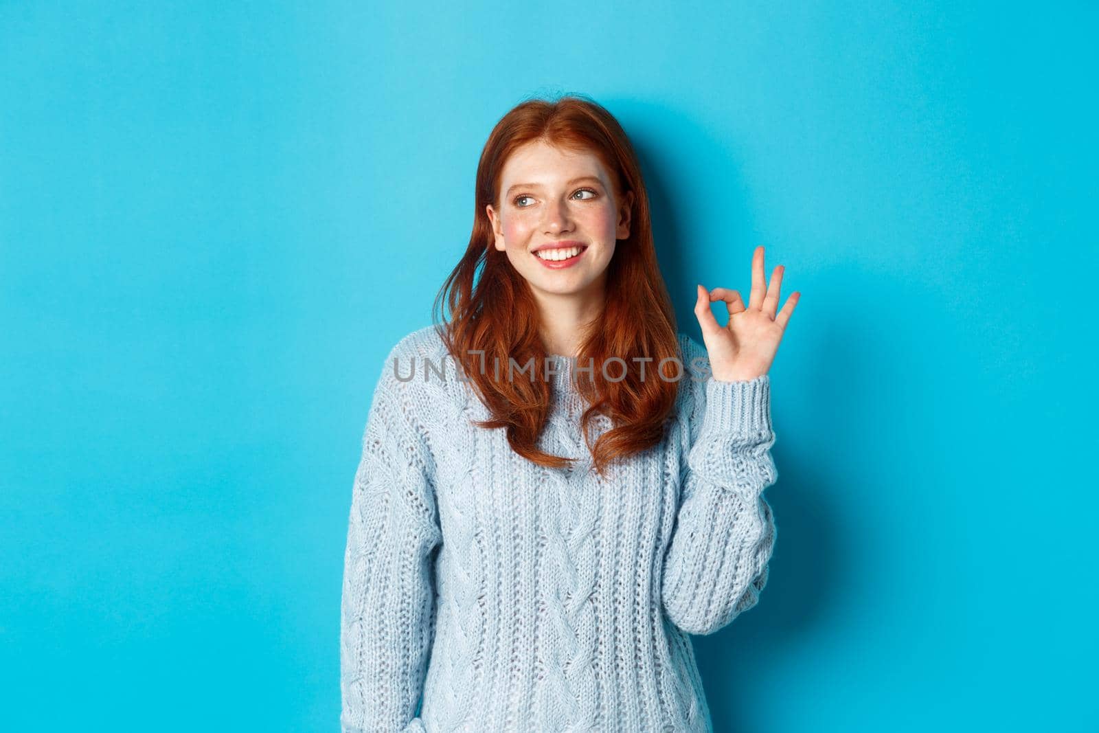 Image of beautiful redhead woman showing okay sign and smiling satisfied, like and agree, standing against blue background.