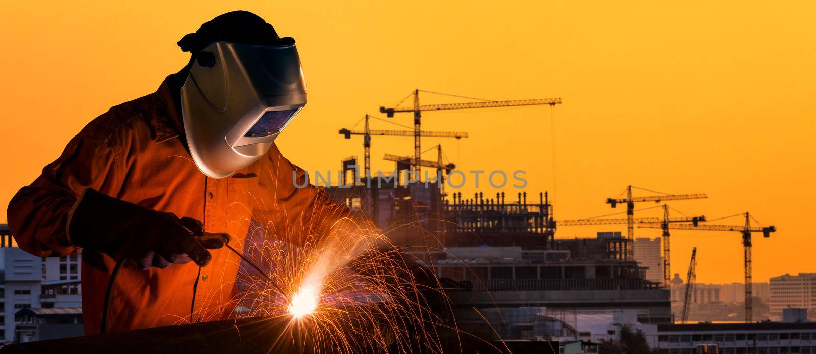 Industrial worker welding steel structure for infrastructure building project with construction site in background.